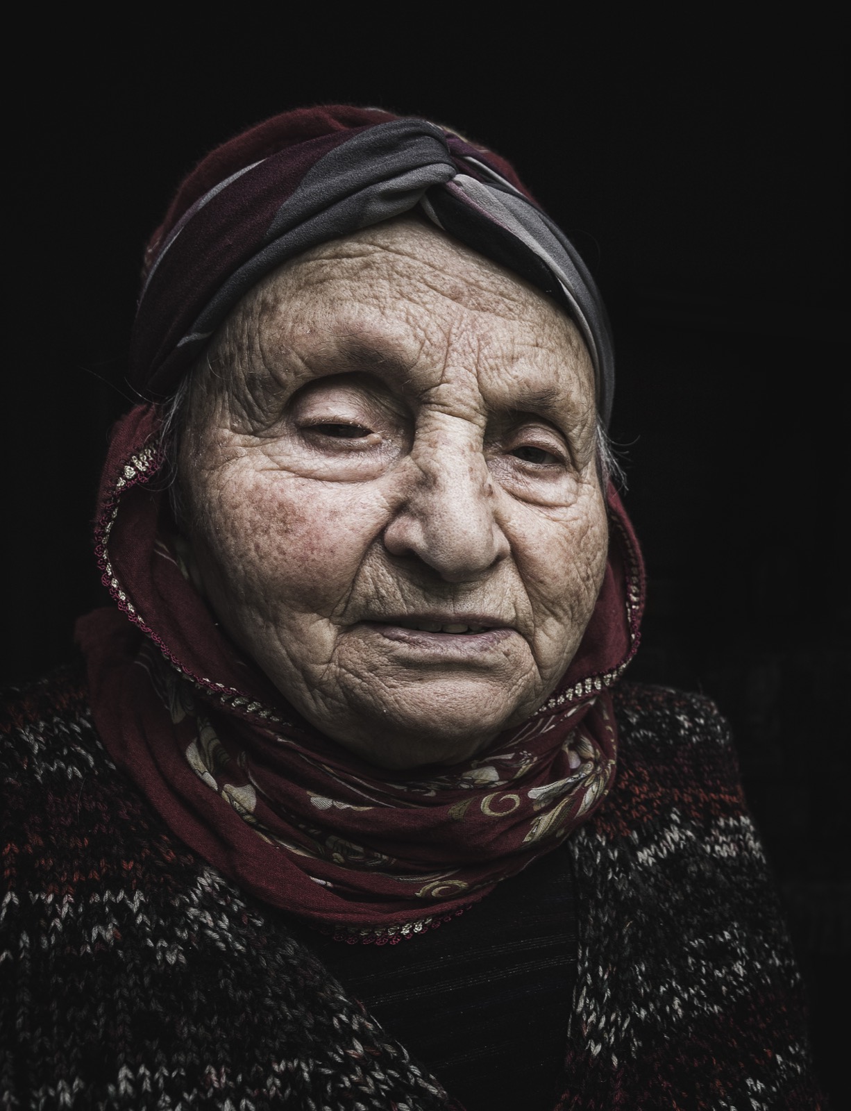 Black and white portrait of an elderly woman in a dark red headscarf and a knitted cardigan, her eyes nearly closed against a deep black background — fine art portrait by Sefa Yamak, Anatolia series.
