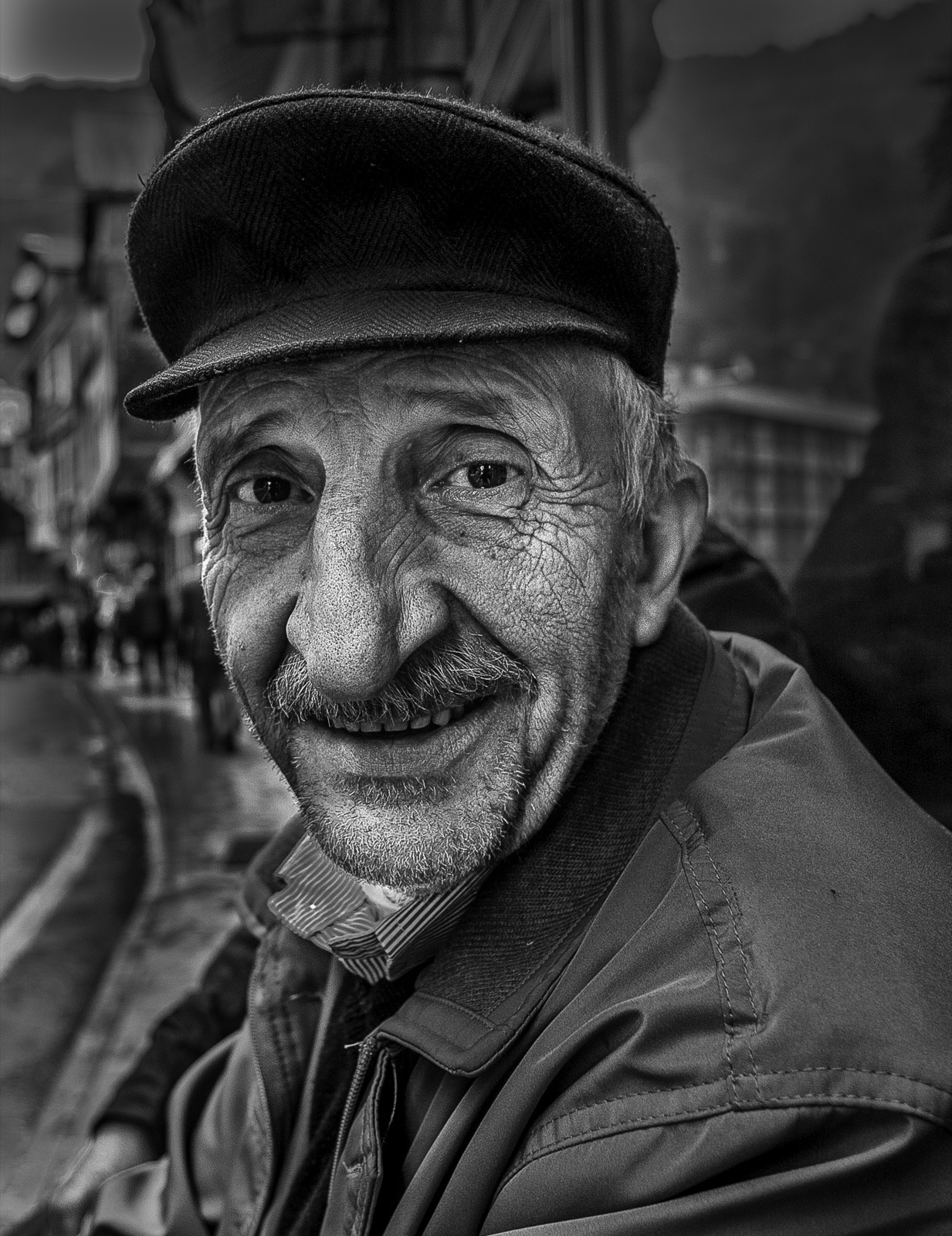 Düz kasketli ve açık renk montlu, gülümseyen yaşlı bir adamın, arkasında insanların silikleştiği ıslak bir kasaba sokağıyla siyah beyaz portresi — Sefa Yamak'tan güzel sanatlar portre, Anadolu serisi.