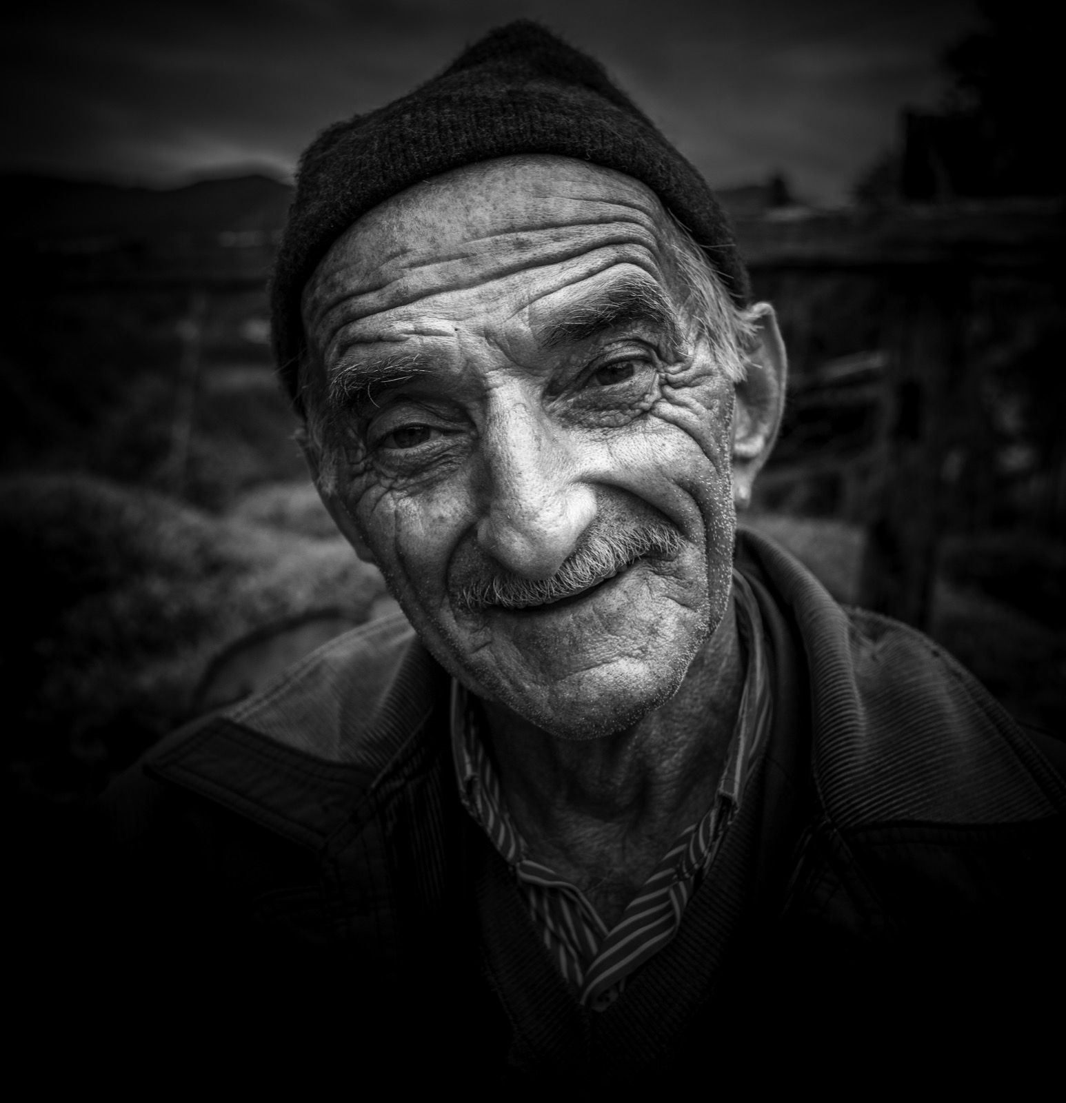 Koyu örgü bere ve çizgili gömlek giyen, yarı gülümseyen yaşlı bir adamın, arkasında karanlık bir yamacın uzandığı siyah beyaz yakın portresi — Sefa Yamak'tan güzel sanatlar portre, Anadolu serisi.