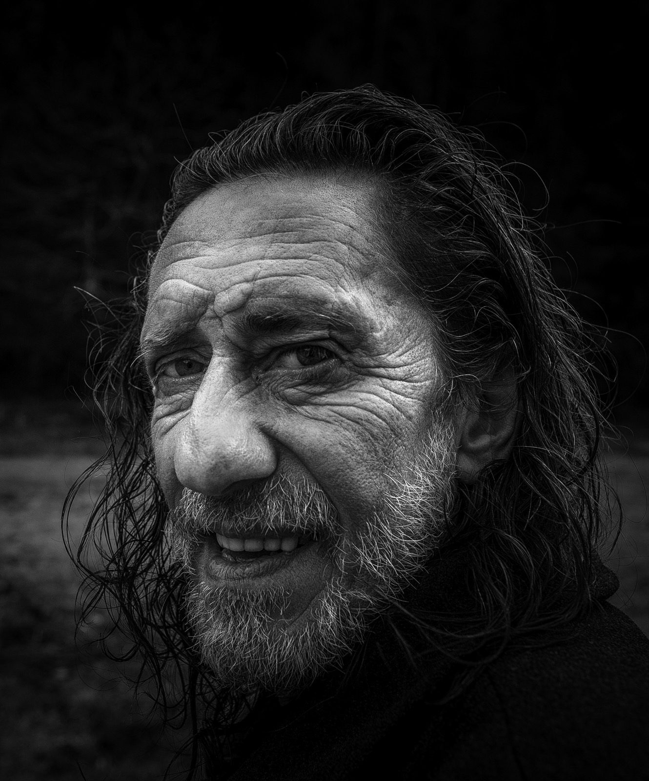 Saçları geriye atılmış, kır karışık sakallı, gülümseyen orta yaşlı bir adamın, arkasında karanlık ormanlık bir fonla siyah beyaz portresi — Sefa Yamak'tan güzel sanatlar portre, Anadolu serisi.