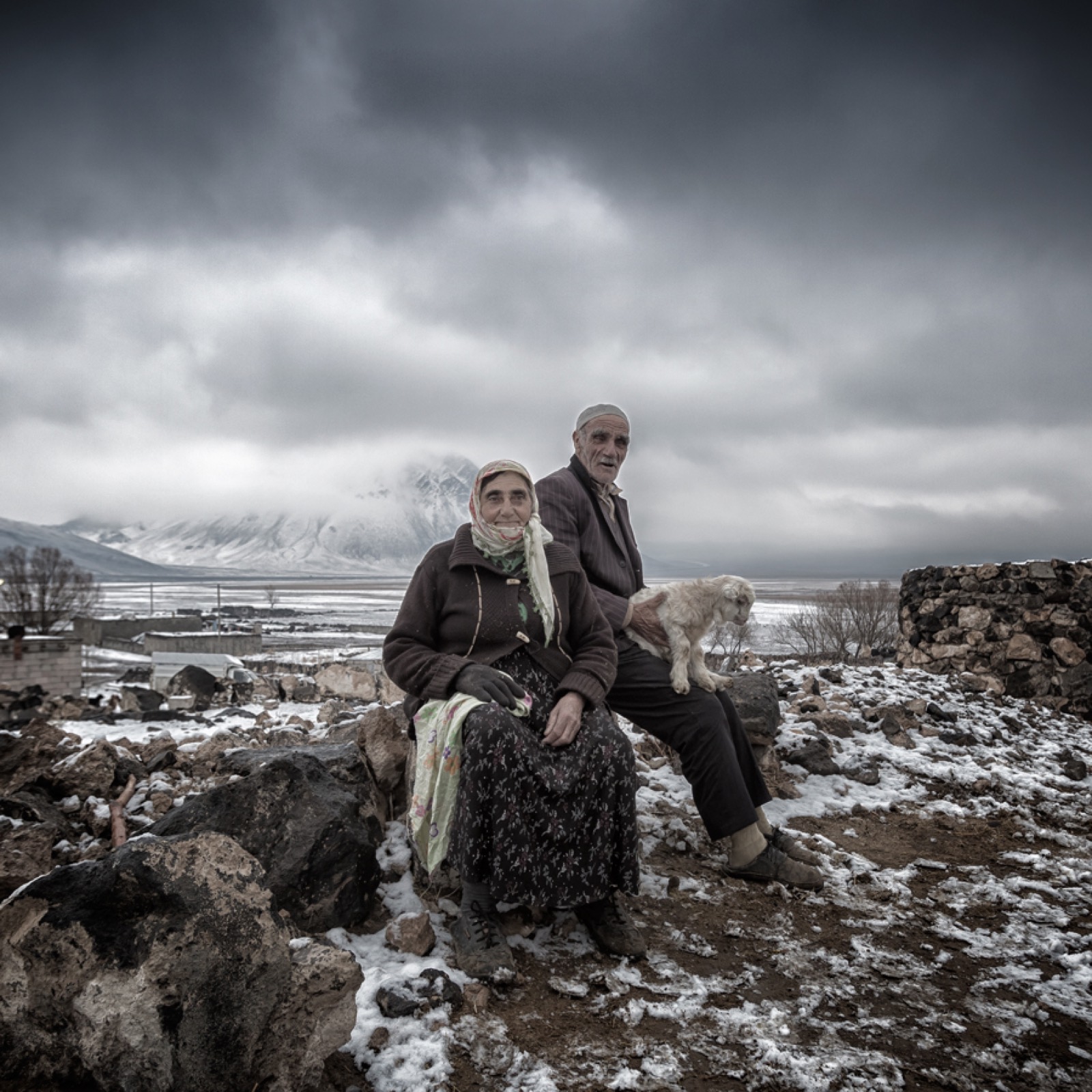 Color portrait of an older couple seated on a stone wall with a small dog, a snowy plain and mountains under heavy cloud behind — fine art portrait by Sefa Yamak, Anatolia series.
