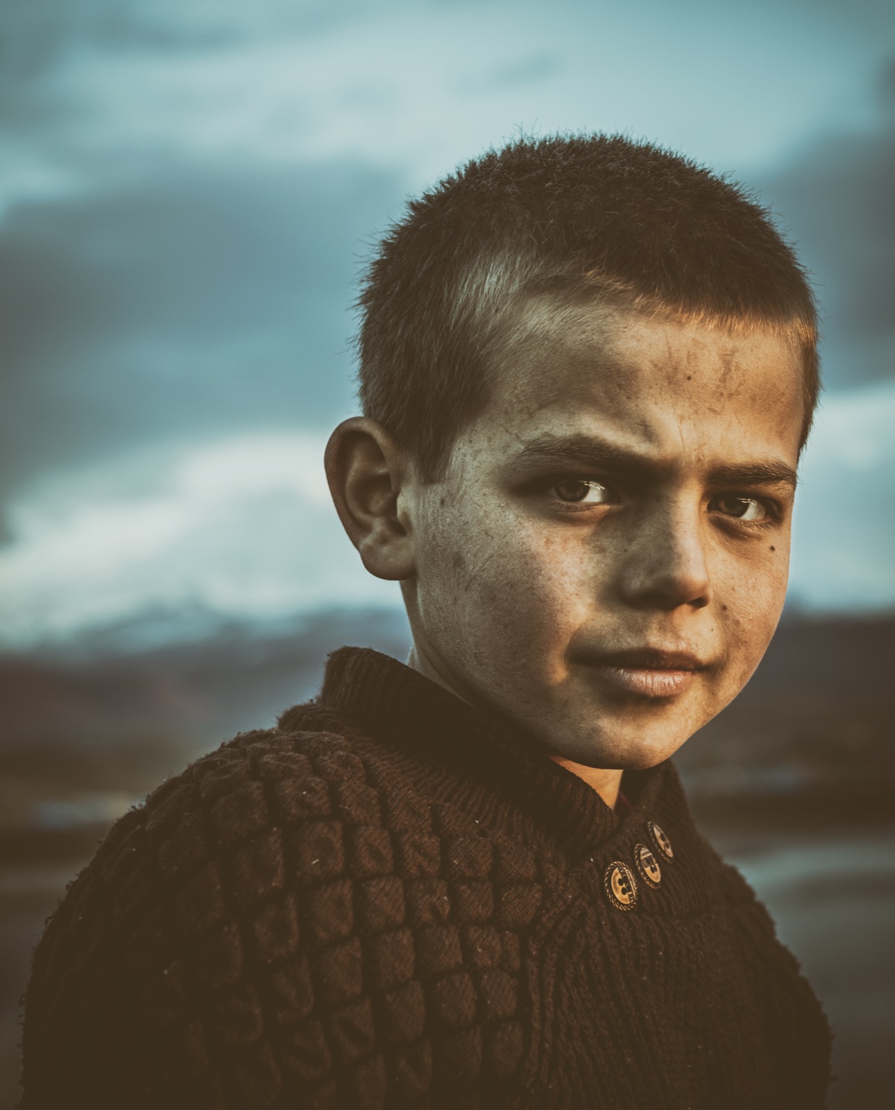 Close portrait of a boy with short hair and a dark knit sweater turning to look back, his face lit warm against a cold blue sky and faint mountains — fine art portrait by Sefa Yamak, Anatolia series.