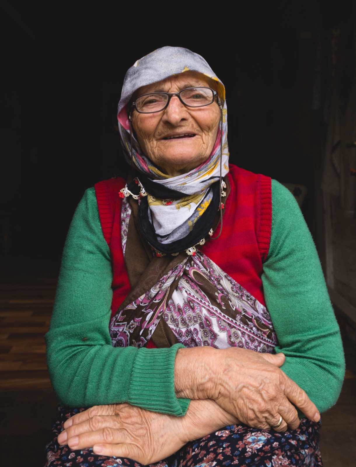 Gözlüklü ve başörtülü, yeşil hırka üzerine kırmızı yelek ve çiçekli önlük giymiş, elleri kucağında kavuşturulmuş, gülümseyen yaşlı bir kadının renkli portresi — Sefa Yamak'tan güzel sanatlar portre, Anadolu serisi.
