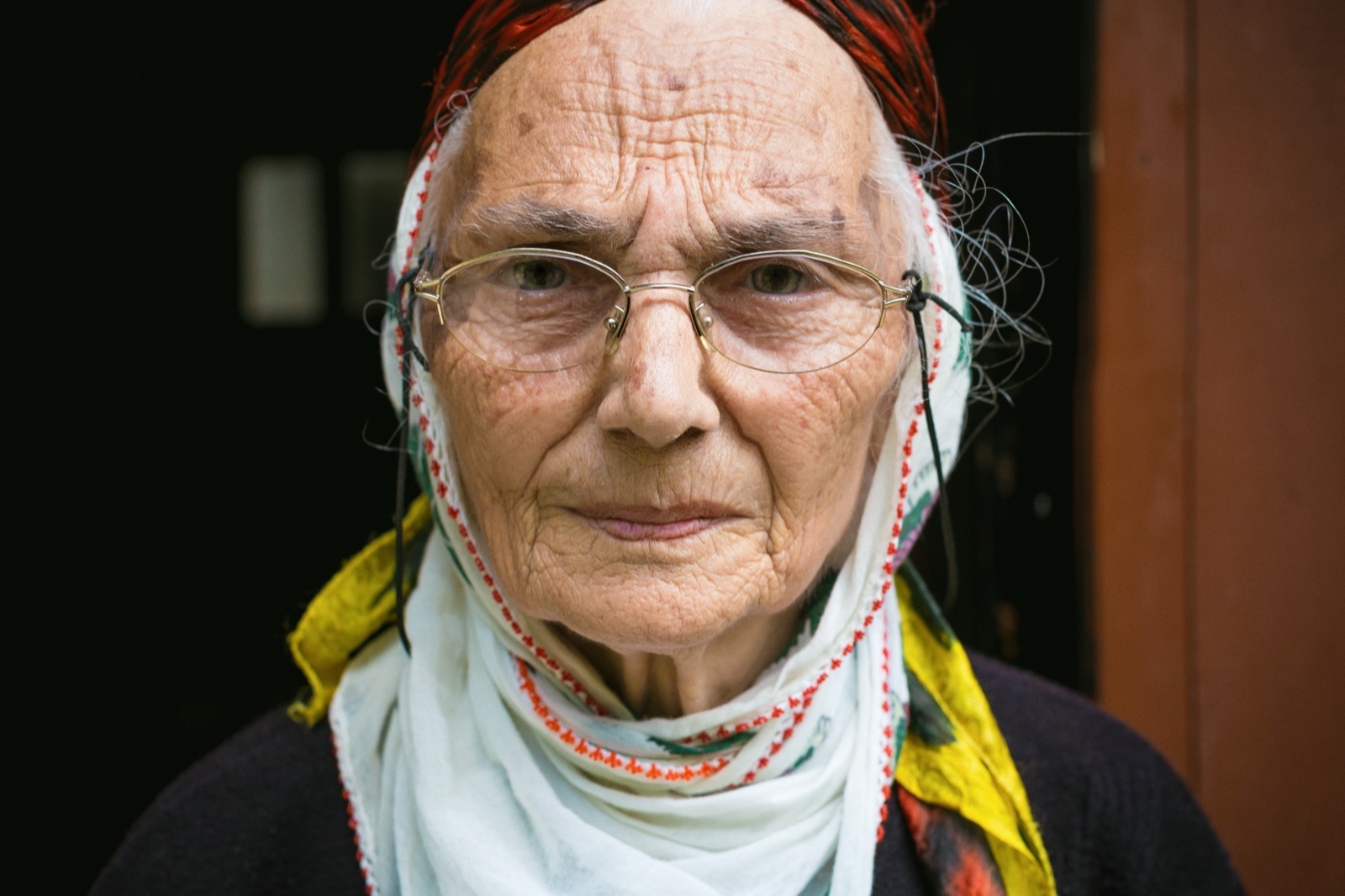 Eski tel çerçeveli gözlük ve kırmızı kenarlı beyaz başörtüsü takan yaşlı bir kadının, bir kapı aralığında doğruca öne baktığı renkli yakın portresi — Sefa Yamak'tan güzel sanatlar portre, Anadolu serisi.