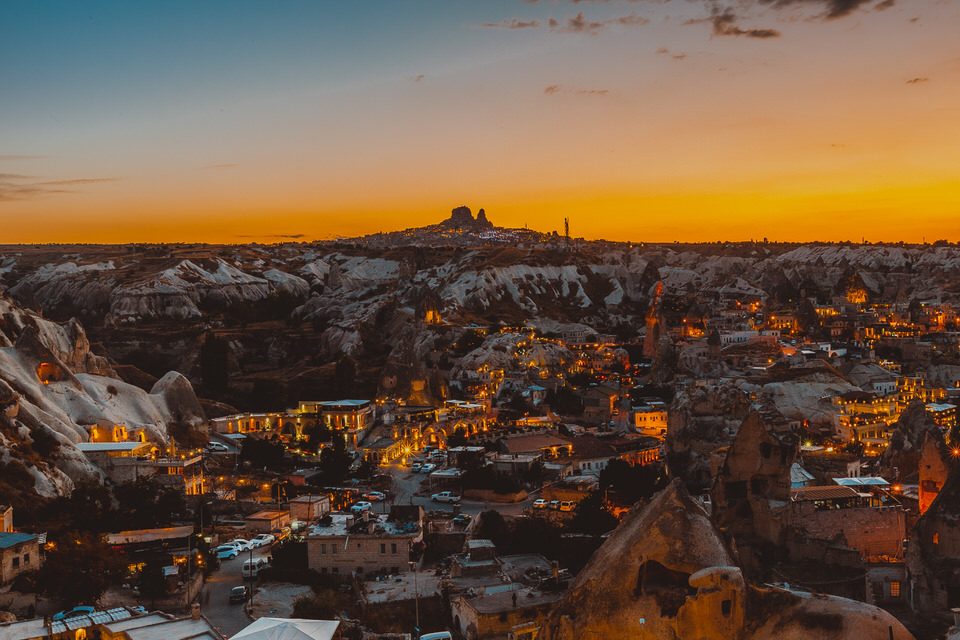 Göreme at dusk seen from above, warm village lights glowing among the rocks, Uçhisar castle on the horizon