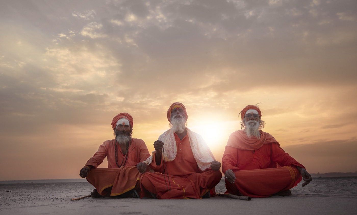 Three sadhus in saffron robes meditate on a riverbed at sunset near Varanasi — fine art portrait by Sefa Yamak, Sadhus series.