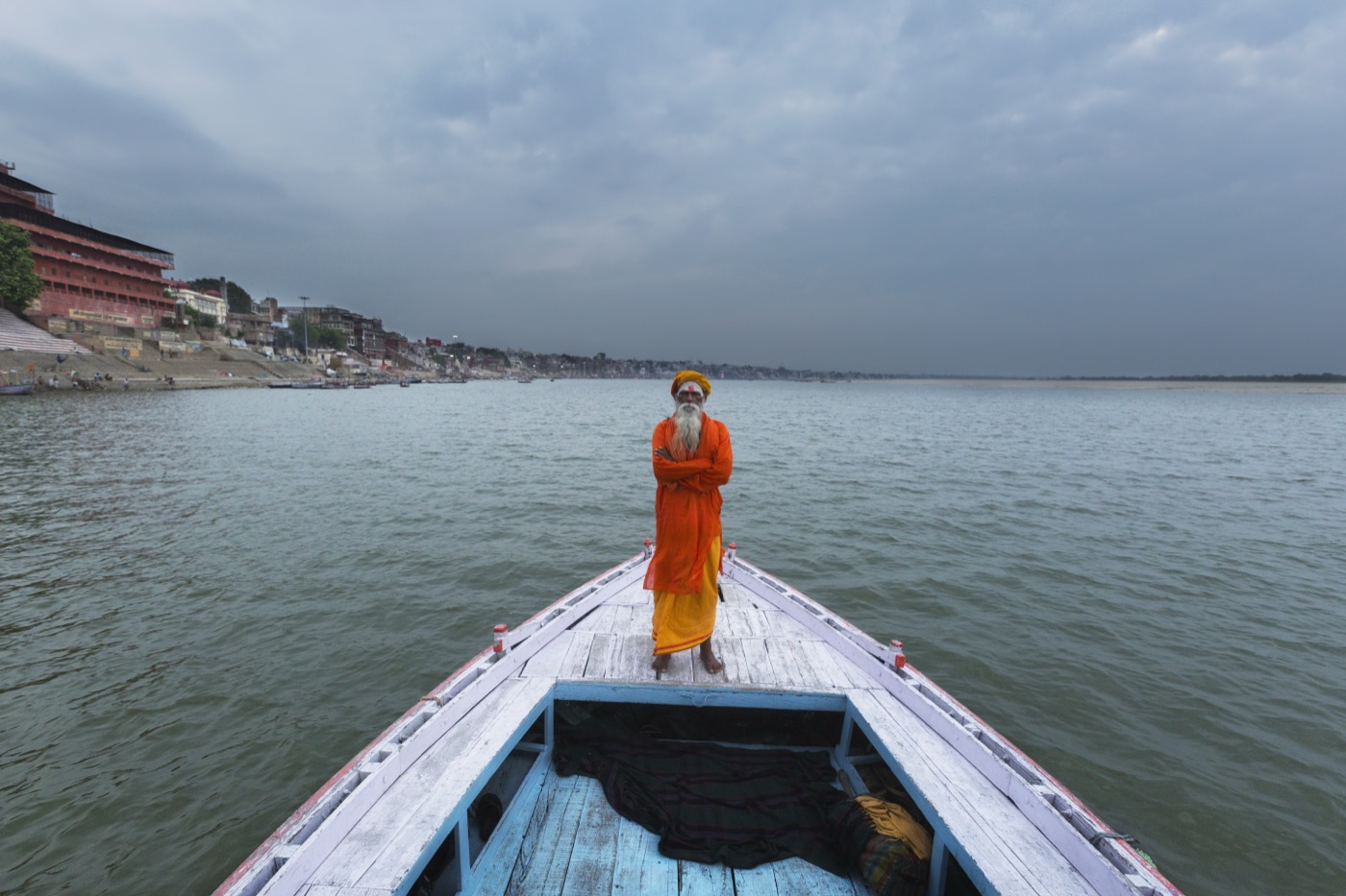 Yalnız bir sadhu, gri gökyüzü altında arkasında Varanasi ghatlarıyla teknenin pruvasında duruyor — Sefa Yamak, Sadhular serisi.