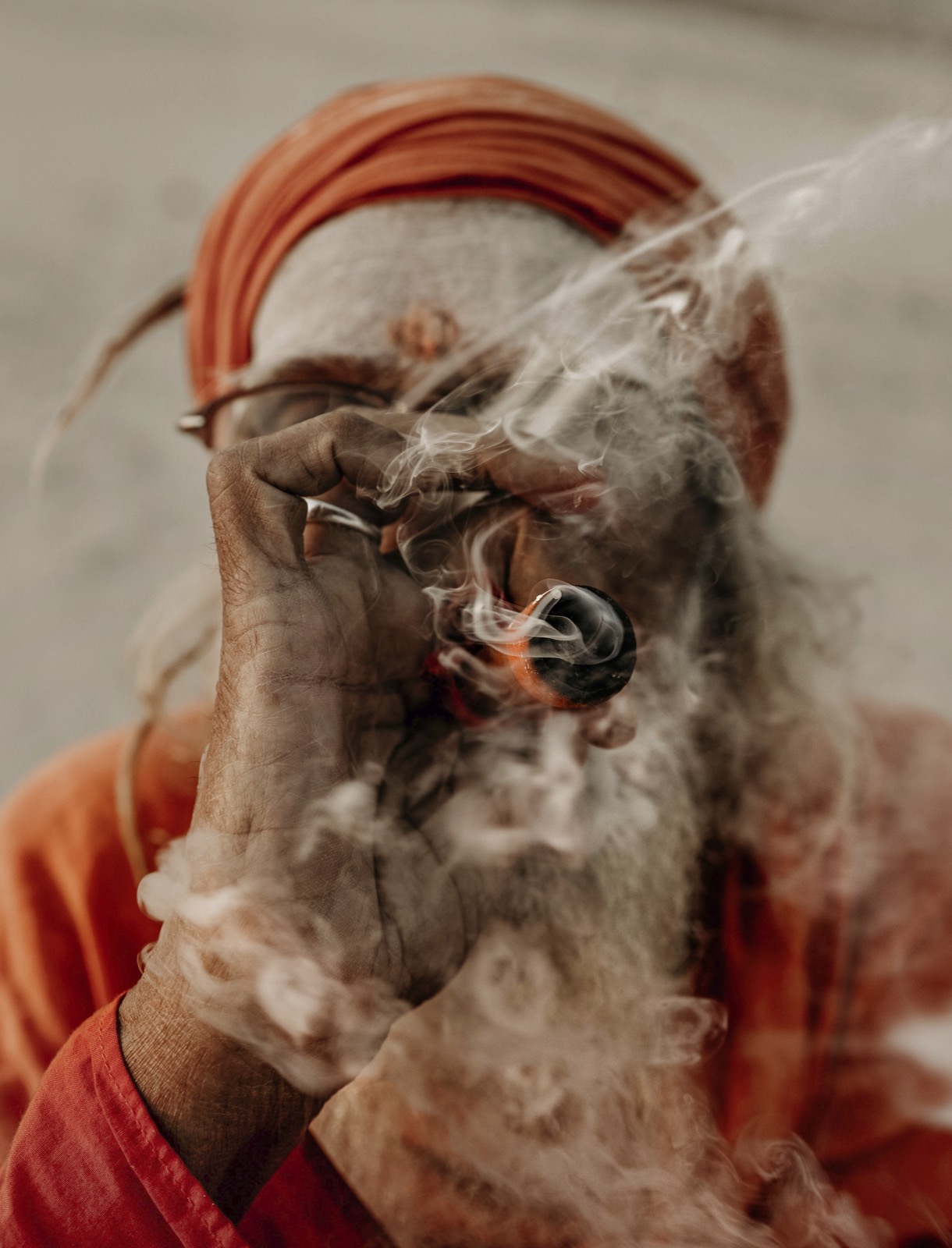 A close view of an elderly man in an orange head wrap drawing on a small clay pipe, his face and long grey beard partly hidden behind a curl of smoke — fine art portrait by Sefa Yamak, India series.