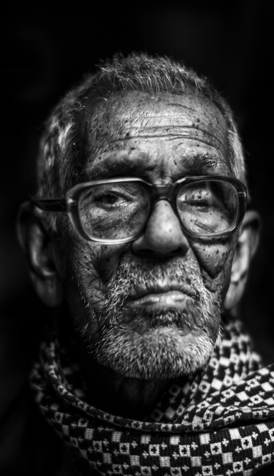 Kalın çerçeveli gözlüklü, desenli atkılı yaşlı bir adamın siyah-beyaz yakın portresi; çizgili yüzü neredeyse tam karanlıktan beliriyor — Sefa Yamak'tan güzel sanatlar portre, Hindistan serisi.