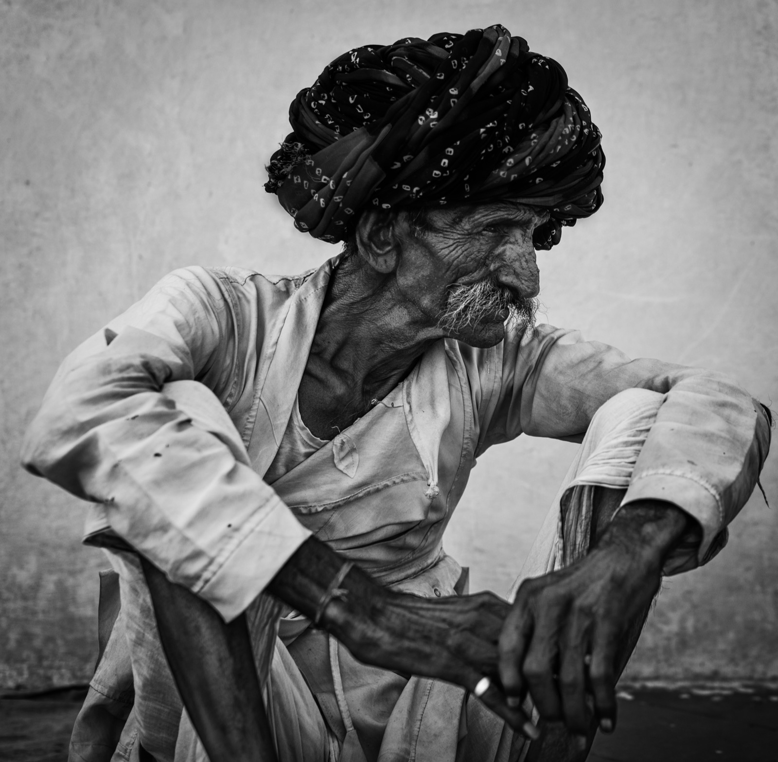 An older man in a patterned turban and worn light clothes sits with his arm resting on his raised knee — fine art portrait by Sefa Yamak, India series.