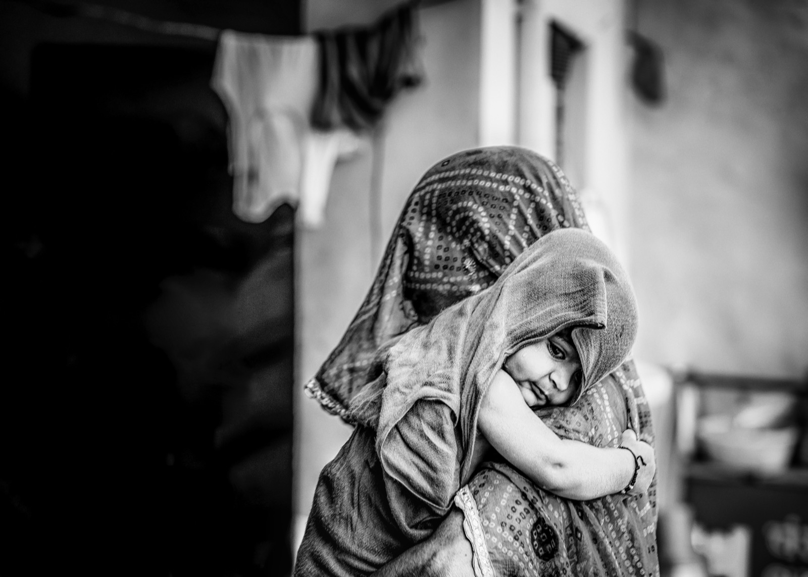 Desenli bir örtüye sarılmış küçük bir çocuk, arkalarında asılı çamaşırlarla bir kadının omzuna yaslanmış tutuluyor — Sefa Yamak'tan güzel sanatlar portre, Hindistan serisi.
