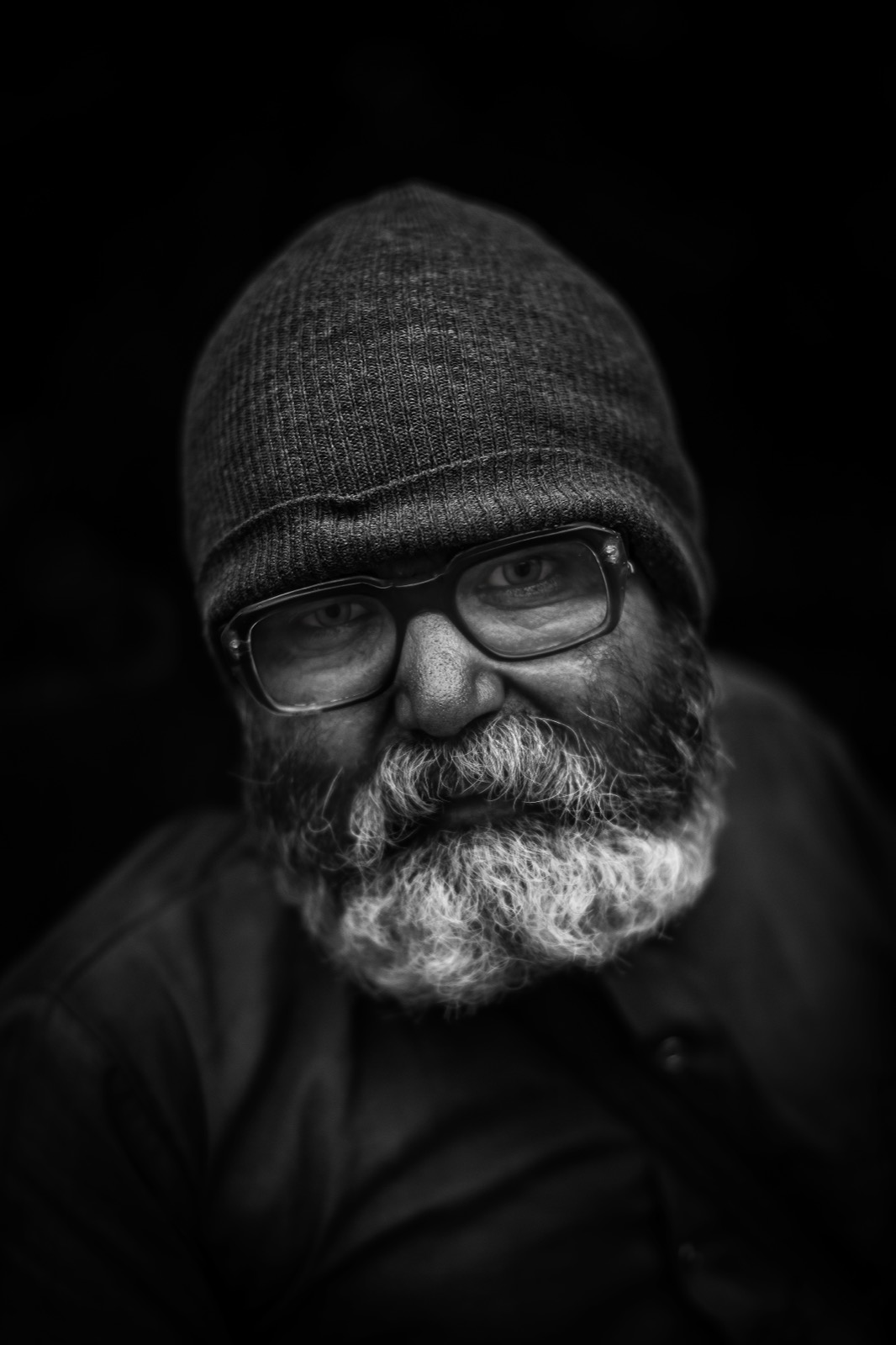 Örgü bere ve kalın çerçeveli gözlük takmış sakallı bir adam, hafifçe gülümseyerek derin gölgeden beliriyor — Sefa Yamak'tan güzel sanatlar portre, Hindistan serisi.