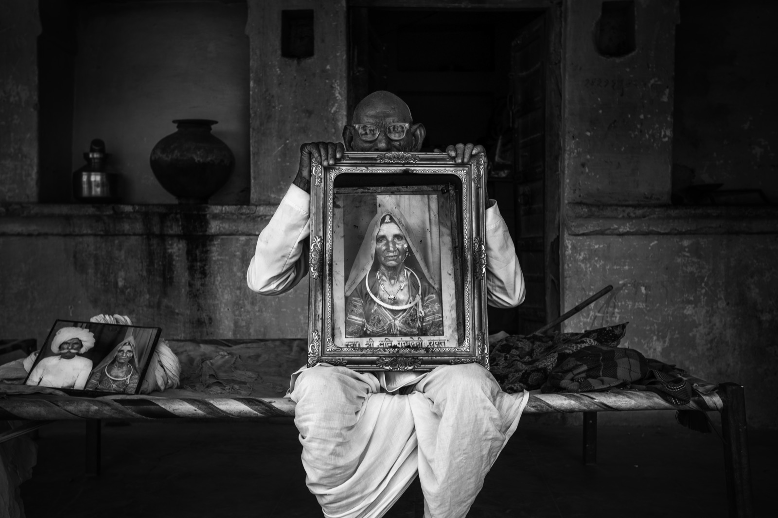 A man in white sits on a cot and holds a large framed portrait of a veiled woman up over his own face in a courtyard — fine art portrait by Sefa Yamak, India series.