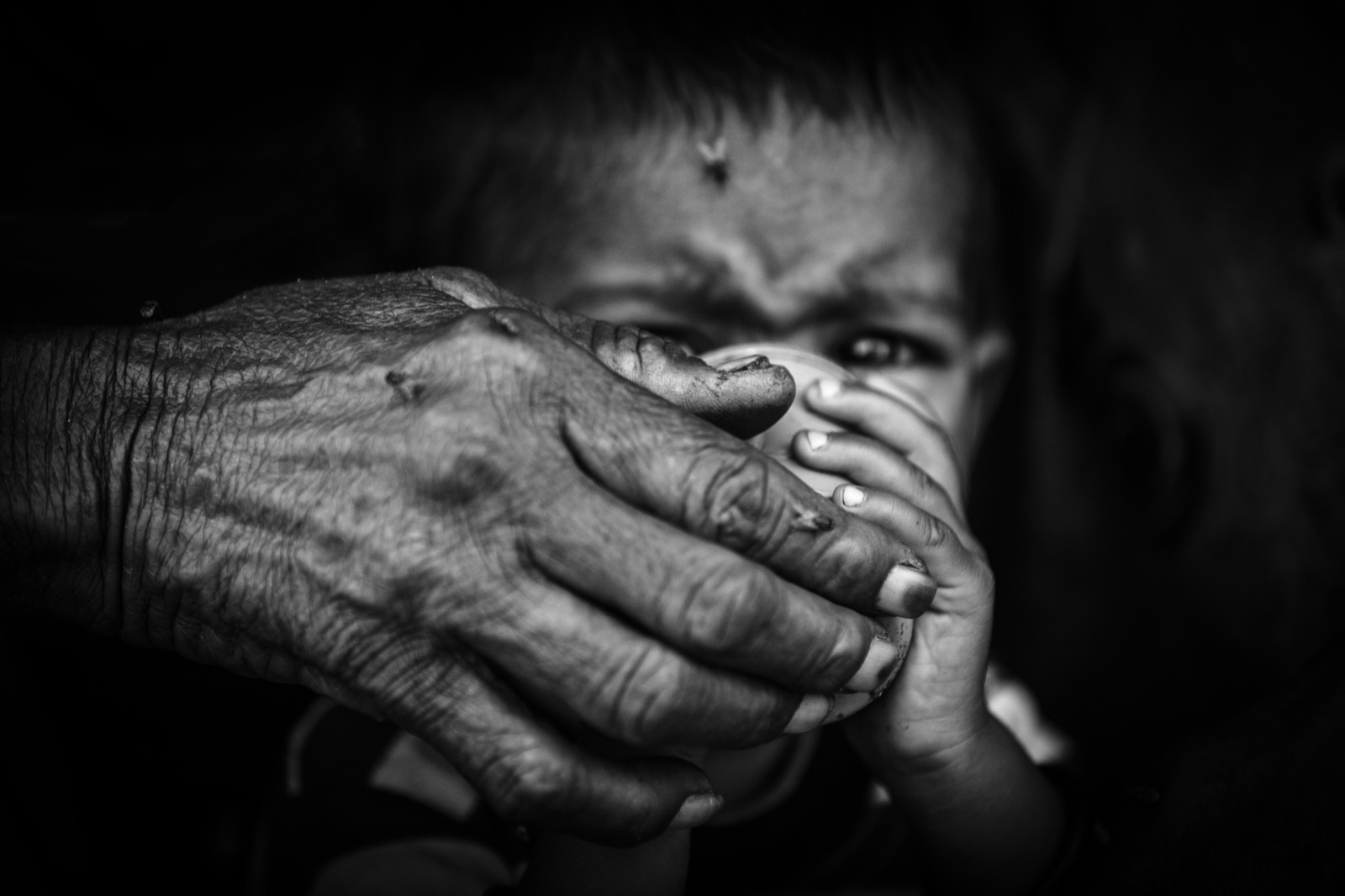 Yaşlı bir yetişkin eli, küçük bir çocuk içerken bardağı ağzına doğru tutuyor, derin gölgeye karşı aydınlatılmış — Sefa Yamak'tan güzel sanatlar portre, Hindistan serisi.