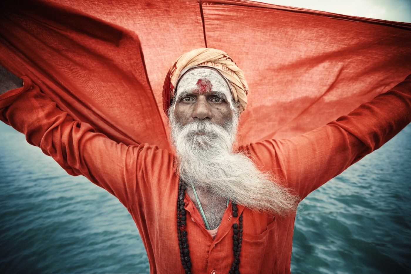 A long-bearded sadhu spreads a red cloth like wings on the Ganges in Varanasi — fine art colour portrait by Sefa Yamak, India series.