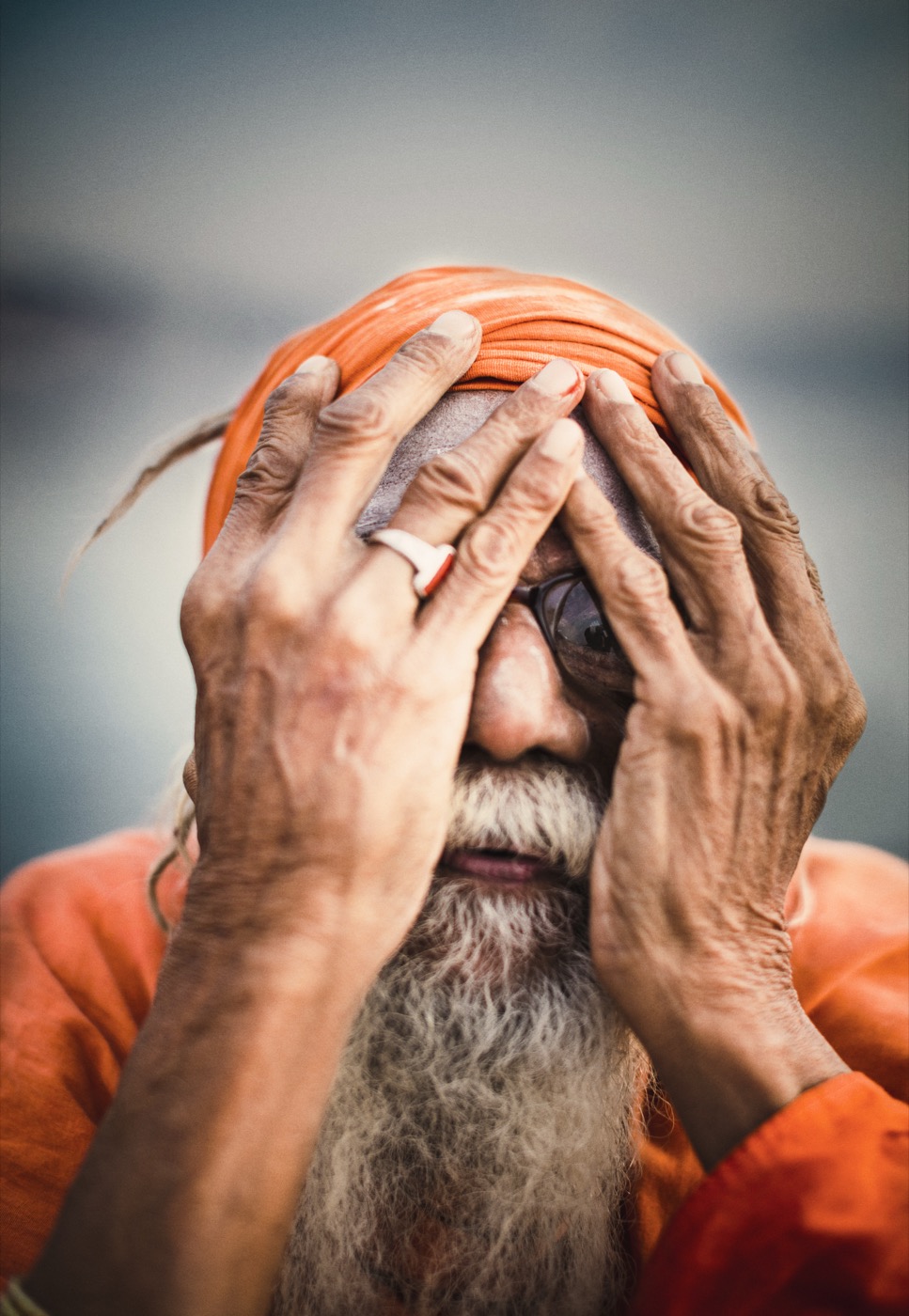 A sadhu hides behind his hands, peeking through his fingers, Varanasi — fine art colour portrait by Sefa Yamak, India series.