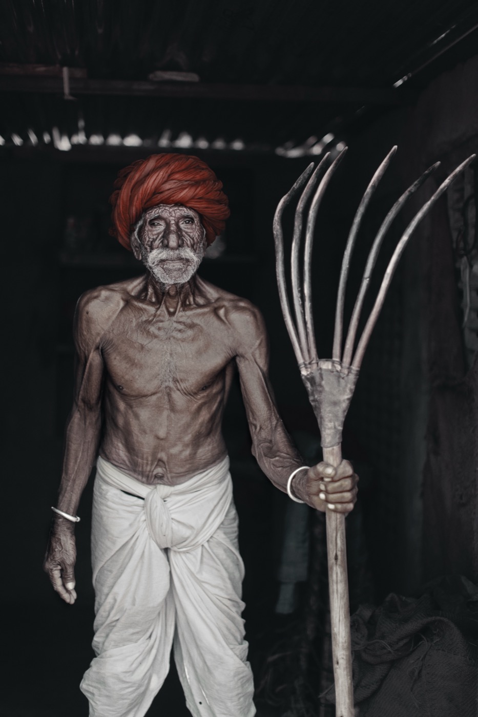A lean farmer in a red Rajasthani turban holds a wooden pitchfork in a dark storeroom in Pushkar — fine art portrait by Sefa Yamak, India series.
