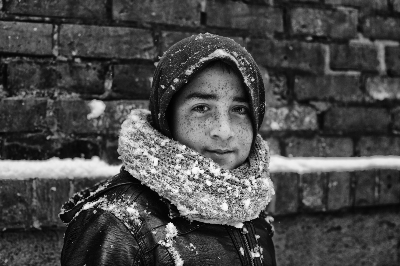 Kapüşonlu ve kalın atkılı, üzerine kar serpilmiş çilli bir çocuk tuğla duvarın önünde duruyor — Sefa Yamak'tan sokak fotoğrafı, İstanbul serisi.
