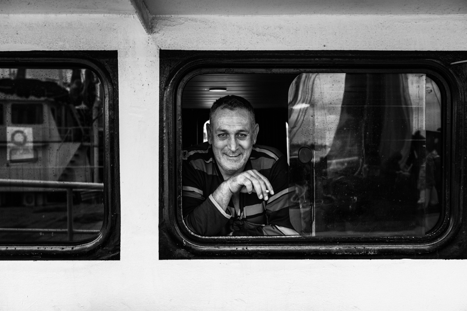 A man leans on the sill inside a ferry window, smiling, with another boat reflected in the glass — street photograph by Sefa Yamak, Istanbul series.