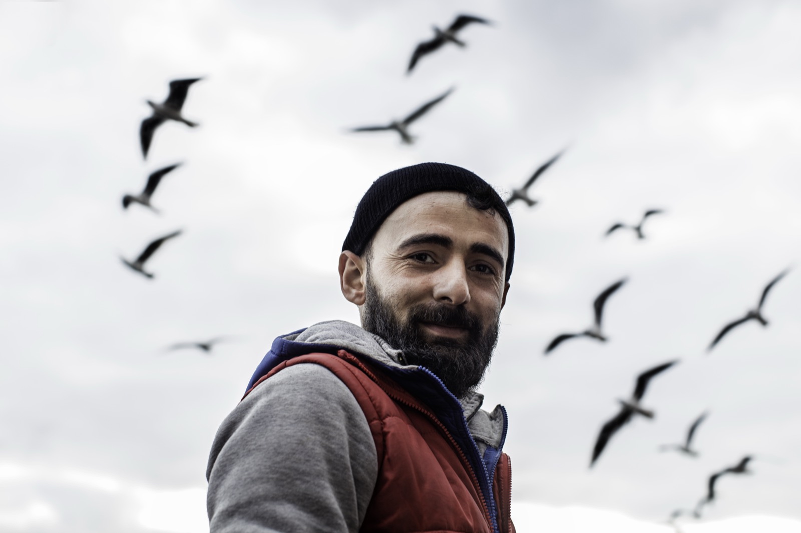 Bereli ve kırmızı yelekli sakallı bir adam, arkasında gri gökyüzünde dönen martılarla gülümsüyor — Sefa Yamak'tan sokak fotoğrafı, İstanbul serisi.