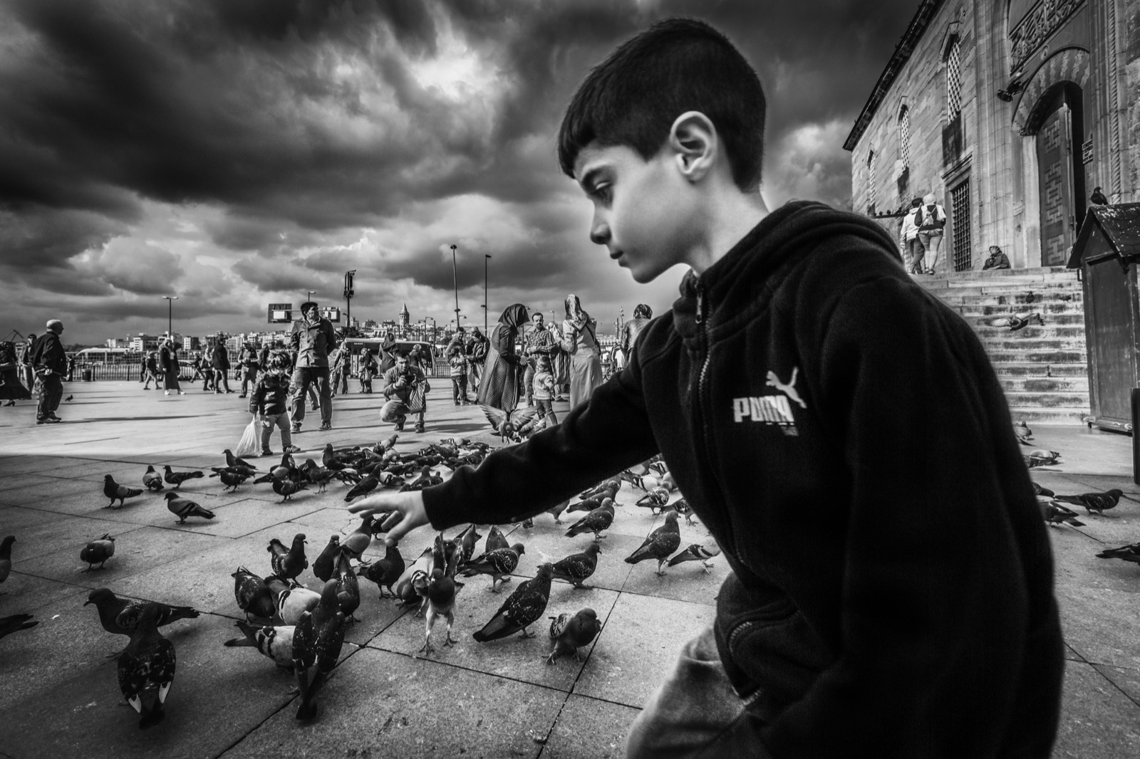 Koyu polarlı bir çocuk ağır bulutların altında cami avlusundaki güvercin sürüsüne elini uzatıyor — Sefa Yamak'tan sokak fotoğrafı, İstanbul serisi.