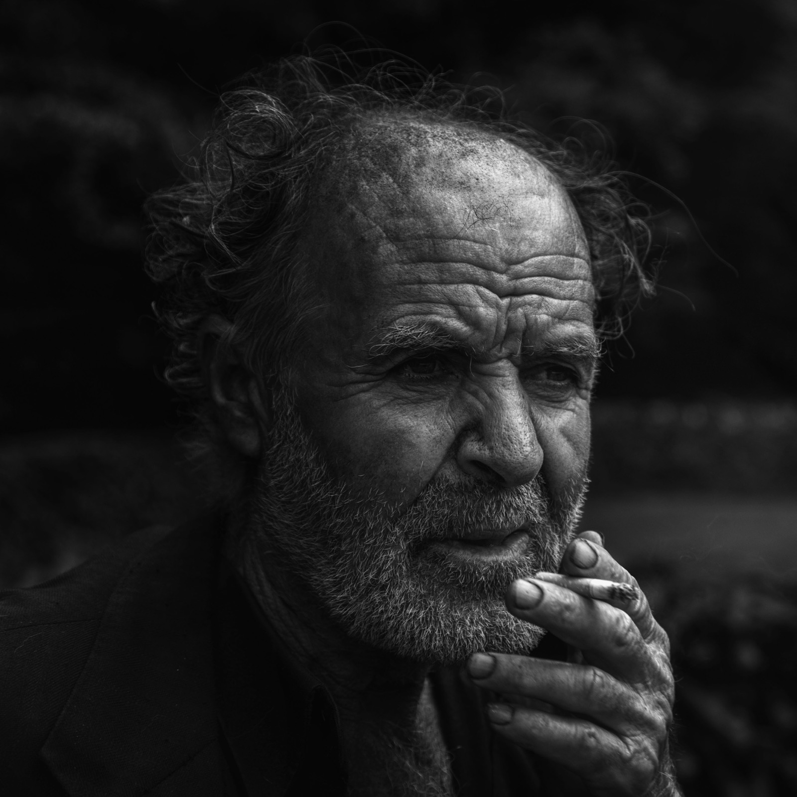 Gri sakallı yaşlı bir adam koyu bir fonun önünde, dalgın bir halde sarma sigara içiyor — Sefa Yamak'tan sokak fotoğrafı, İstanbul serisi.