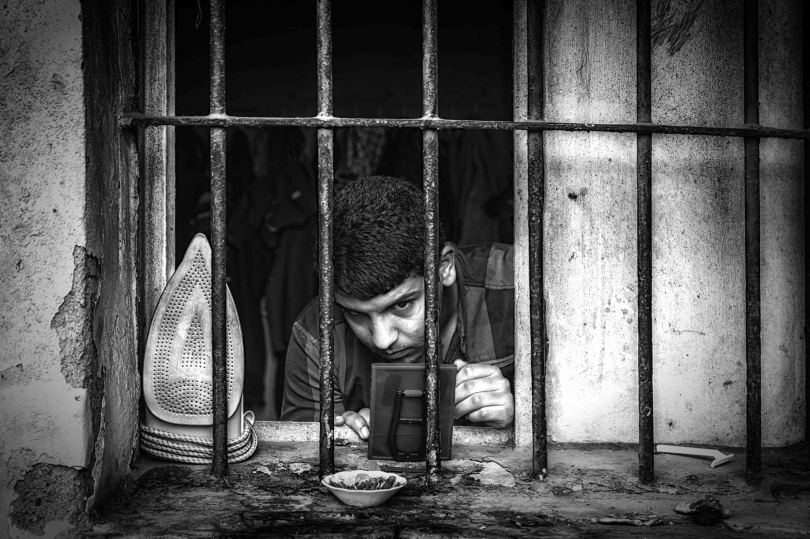 Genç bir adam demir parmaklıkların ardındaki pencere kenarına, eski bir ütü ve küçük bir kâsenin yanına yaslanmış — Sefa Yamak'tan sokak fotoğrafı, İstanbul serisi.