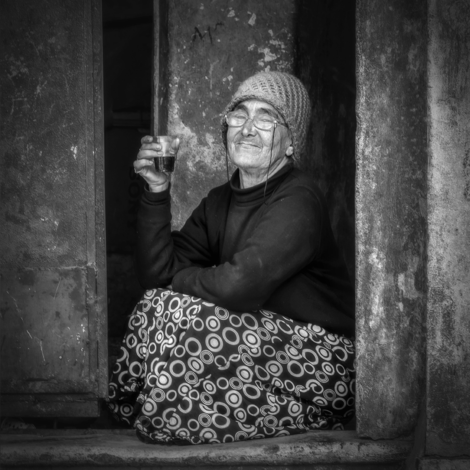 Örgü bereli ve desenli şalvarlı yaşlı bir kadın bir kapı eşiğinde oturmuş elinde bir bardak çay tutuyor — Sefa Yamak'tan sokak fotoğrafı, İstanbul serisi.