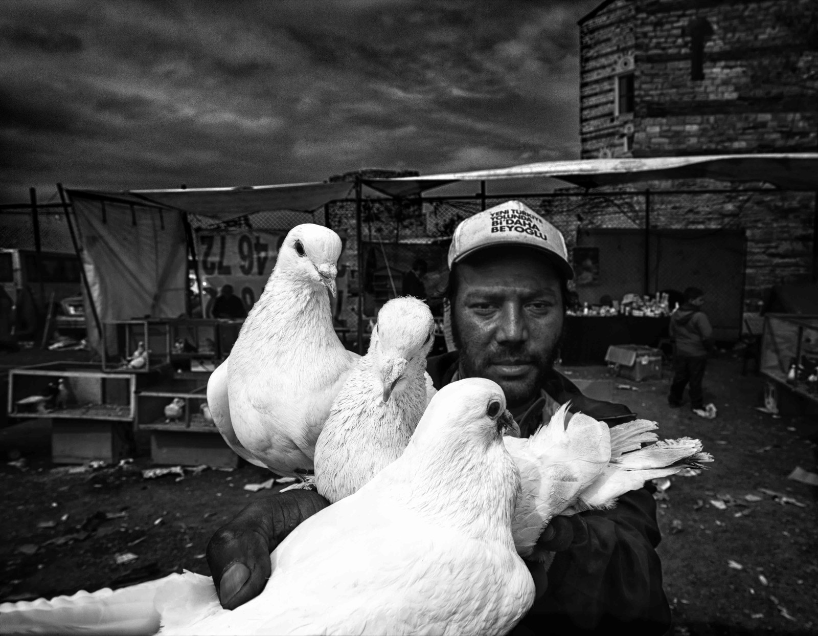 Kasketli bir adam fırtınalı bir gökyüzünün altındaki açık pazarda göğsüne yakın birkaç beyaz güvercin tutuyor — Sefa Yamak'tan sokak fotoğrafı, İstanbul serisi.