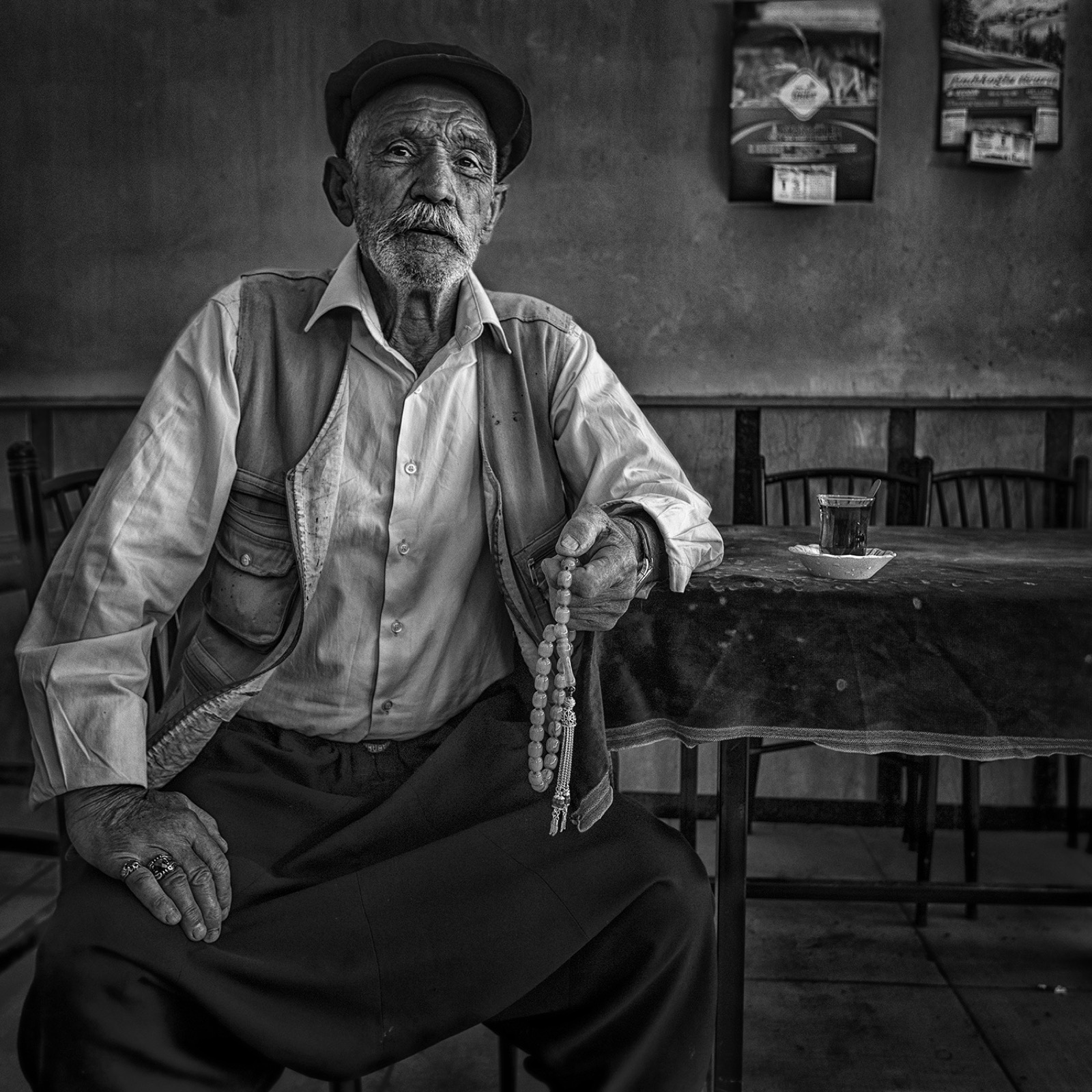 Kasketli ve yelekli yaşlı bir adam çayhanede tespih tutarak oturuyor, yanındaki masada bir bardak çay var — Sefa Yamak'tan sokak fotoğrafı, İstanbul serisi.