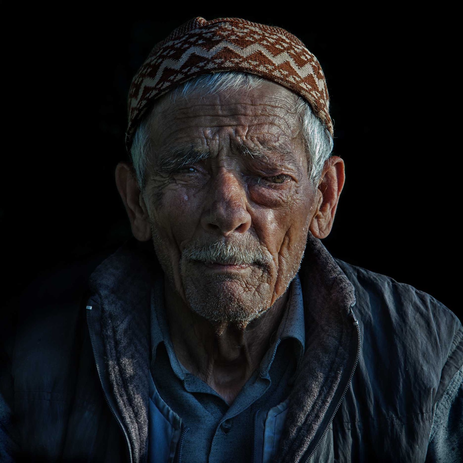 Desenli örgü takkeli ve polar yelekli yaşlı bir adam siyah bir fonun önünde aydınlanmış halde yana bakıyor — Sefa Yamak'tan sokak fotoğrafı, İstanbul serisi.