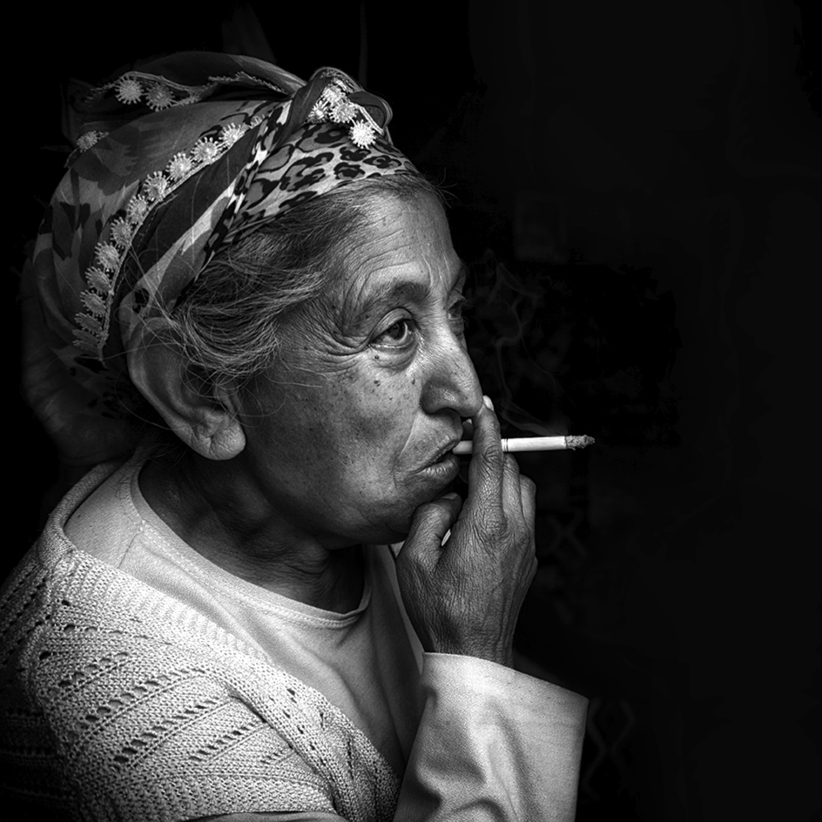 Desenli yazmalı yaşlı bir kadın sigara içerek karanlığa doğru bakıyor — Sefa Yamak'tan sokak fotoğrafı, İstanbul serisi.