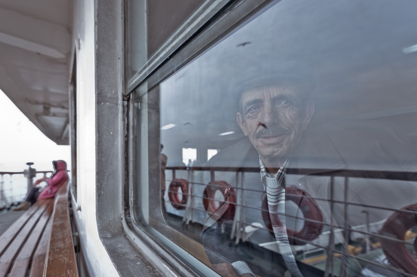 Şapkalı yaşlı bir adam vapur penceresinin ardında oturuyor, camda can simitleri ve güverte yansıyor — Sefa Yamak'tan sokak fotoğrafı, İstanbul serisi.