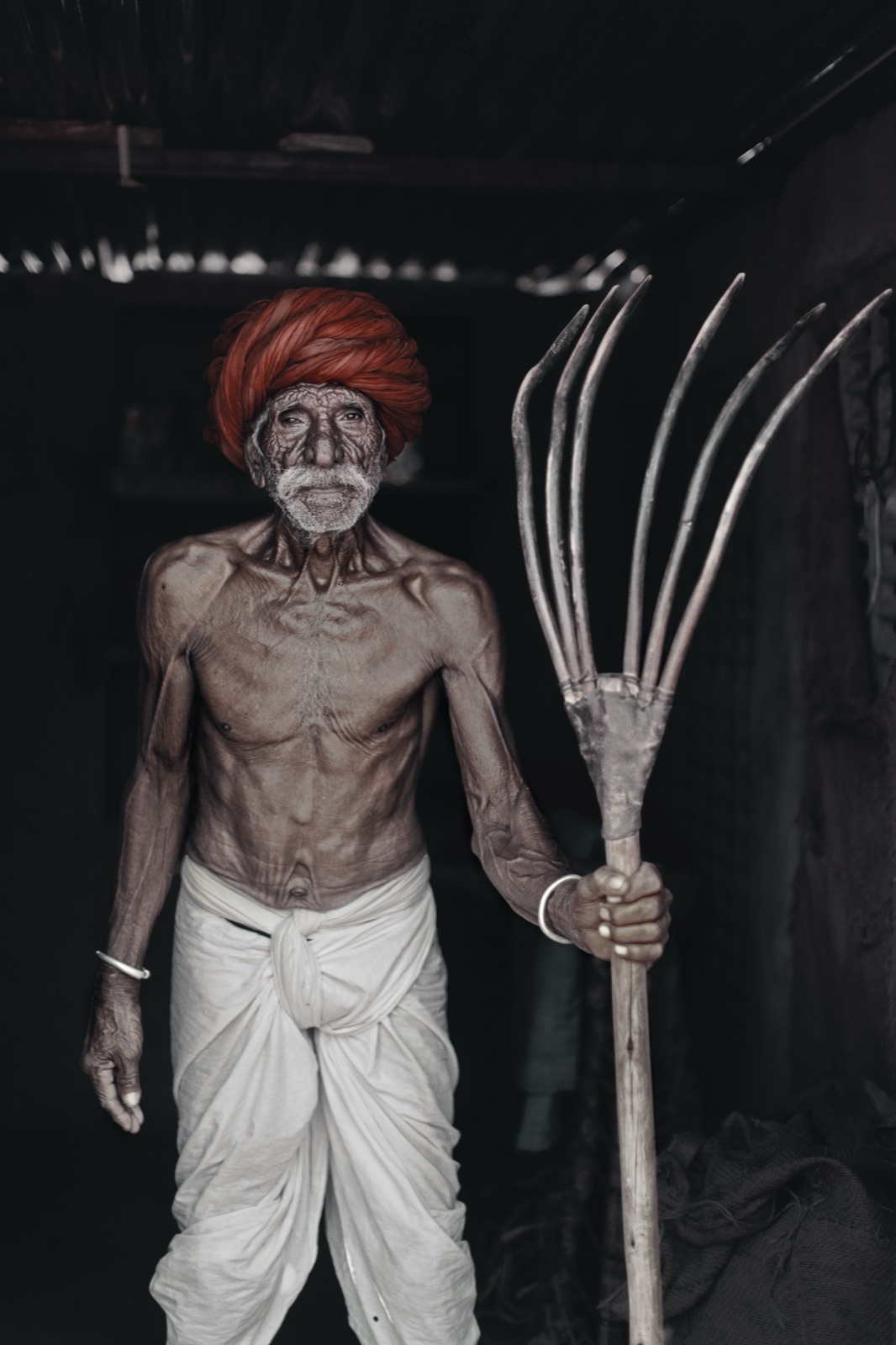 A lean farmer in a red Rajasthani turban holds a wooden pitchfork in a dark storeroom in Pushkar — fine art portrait by Sefa Yamak, India series.