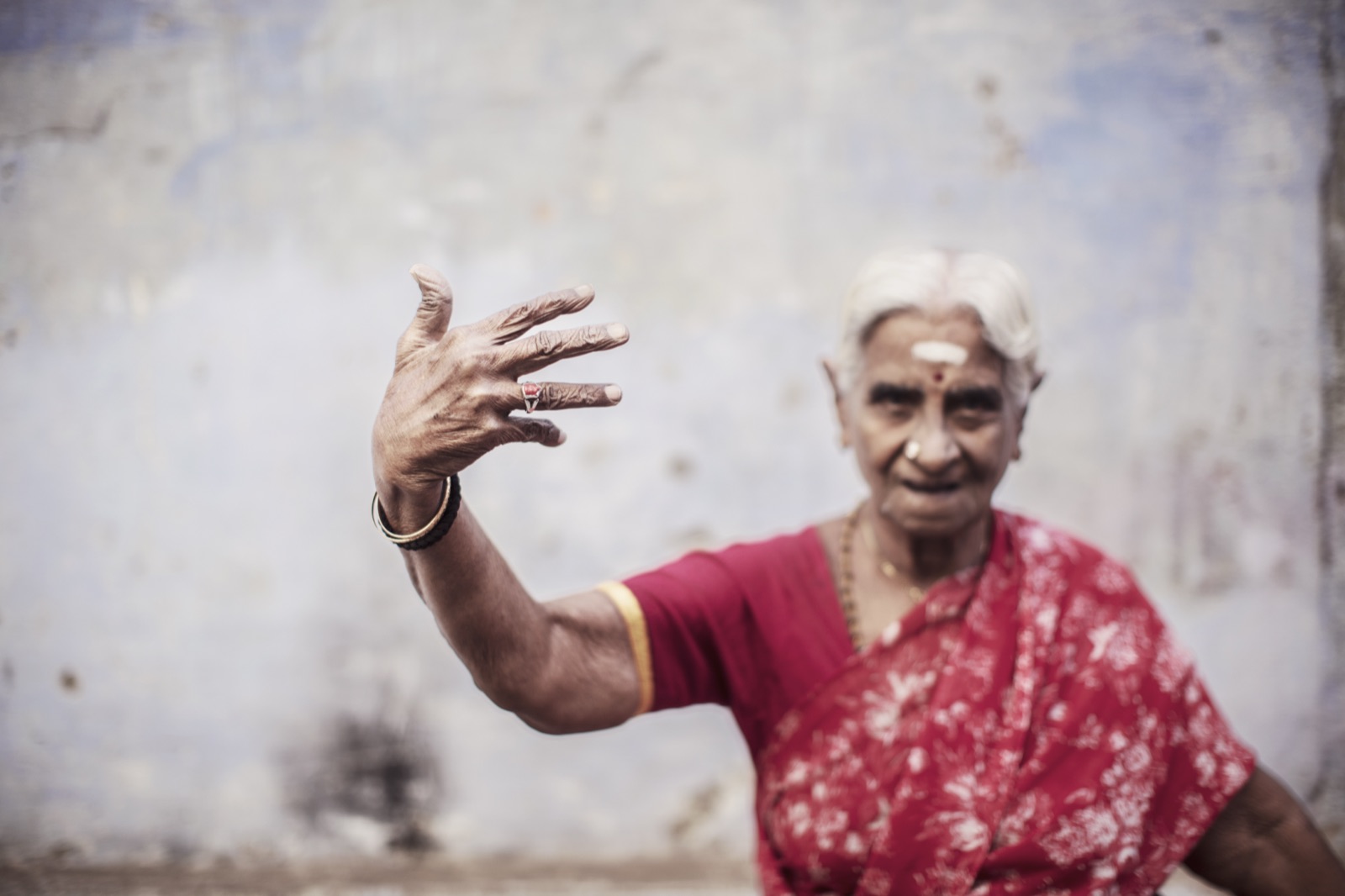 An elderly woman in a red sari raises an open hand toward the camera, southern India — fine art colour portrait by Sefa Yamak, India series.