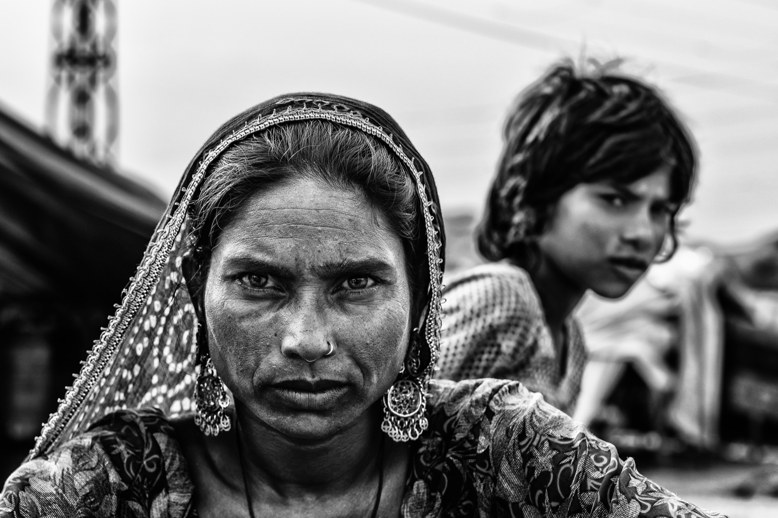 A woman with silver earrings stares into the lens as a child watches behind her in Pushkar — fine art black-and-white portrait by Sefa Yamak, India series.
