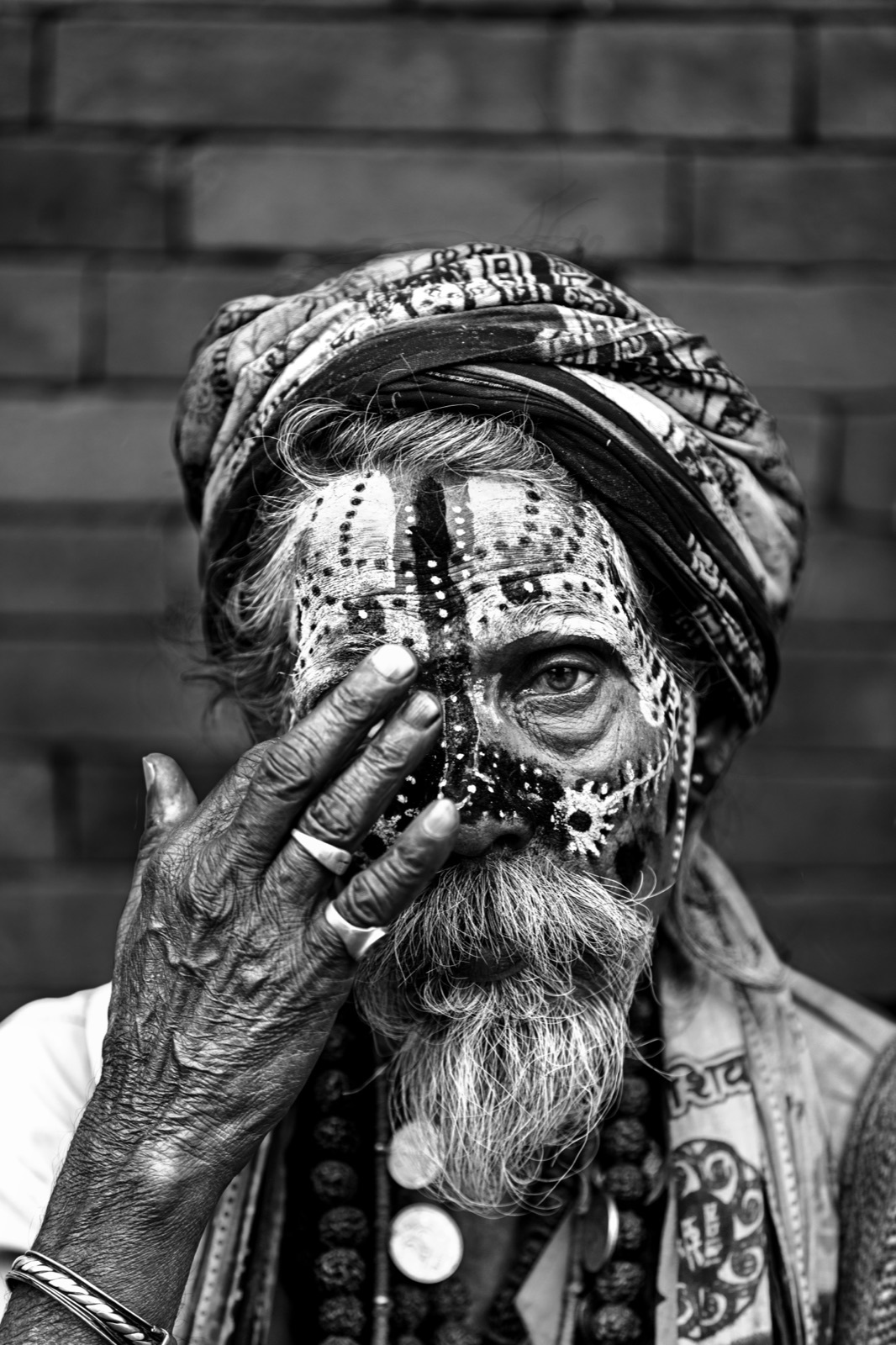 A sadhu with dotted face paint lifts a hand to his eye against a brick wall in Varanasi — fine art black-and-white portrait by Sefa Yamak, India series.