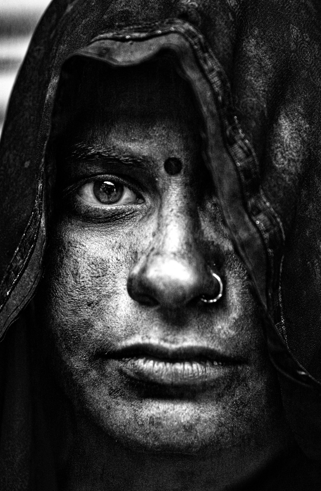 A woman's face half-lit inside a dark hood, one eye meeting the lens, Mathura — fine art black-and-white portrait by Sefa Yamak, India series.