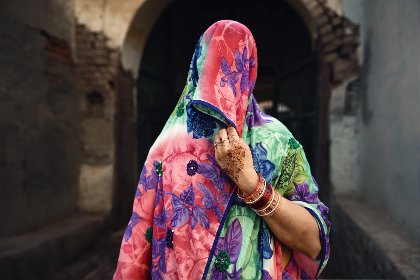 A woman draws a floral veil across her face, henna on her hand, by a stone archway in Rajasthan — fine art colour portrait by Sefa Yamak, India series.