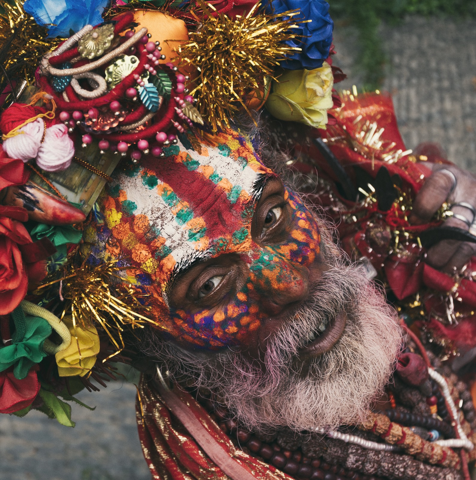Close color portrait, framed at a tilt, of a man with an intricately painted face in orange, green and white, surrounded by artificial flowers and beads — fine art portrait by Sefa Yamak, Nepal series.
