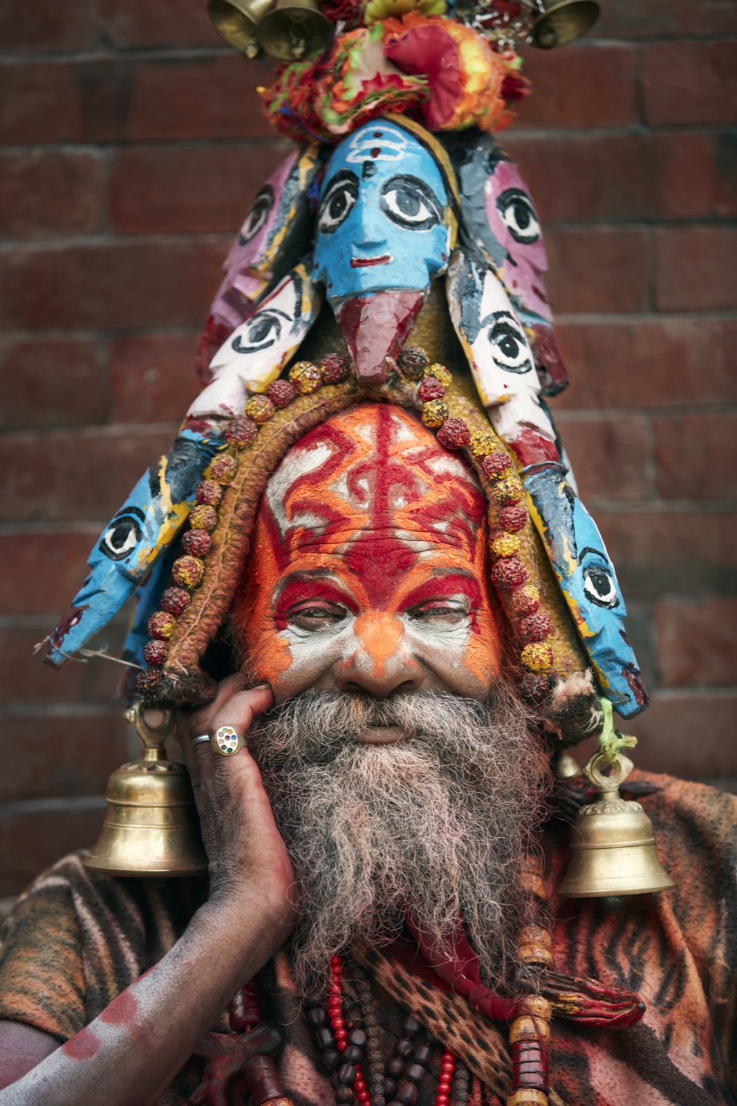 Color portrait of a bearded man with an orange and white painted face wearing a tall headdress of painted deity faces and bells, against a brick wall — fine art portrait by Sefa Yamak, Nepal series.