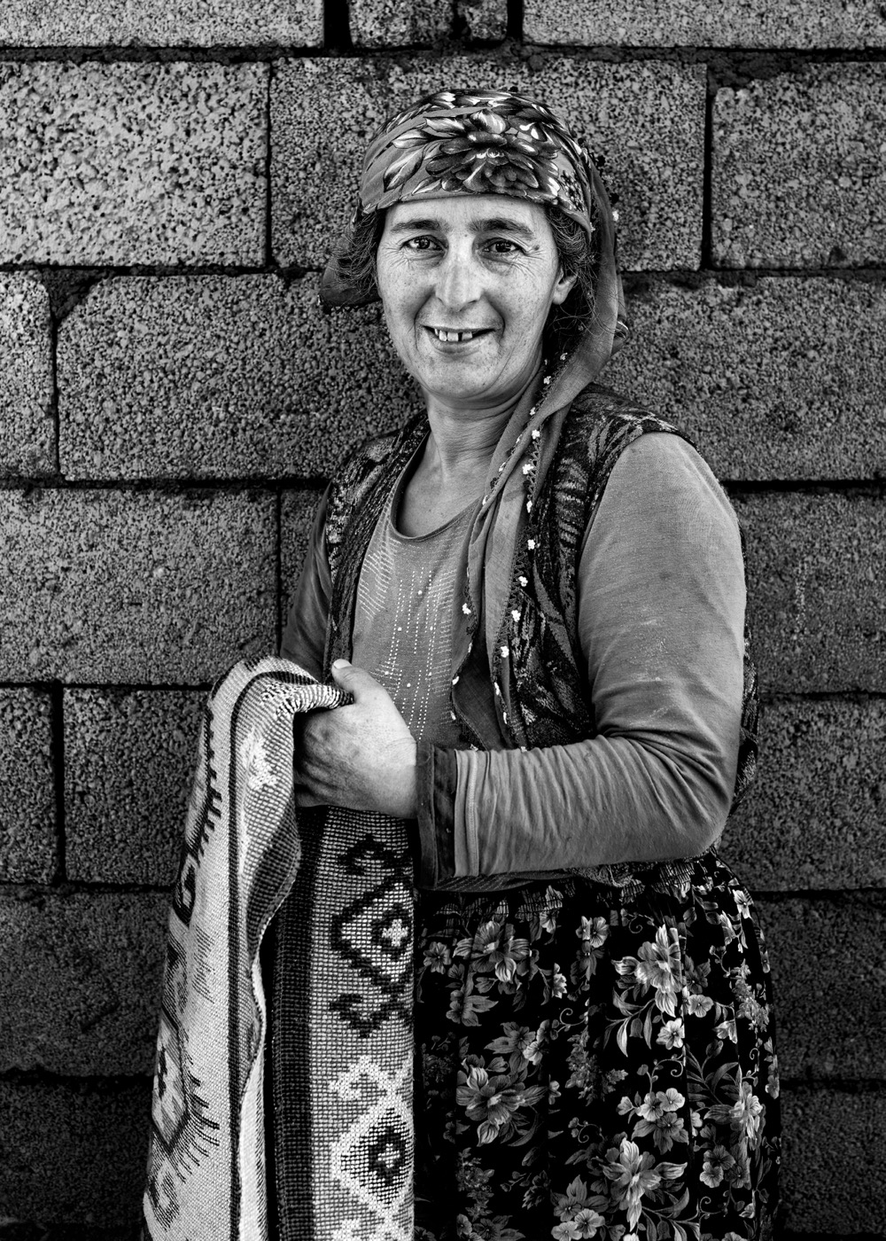 A village woman holds a hand-woven kilim against a breeze-block wall, eastern Anatolia — fine art black-and-white portrait by Sefa Yamak, Anatolia series.