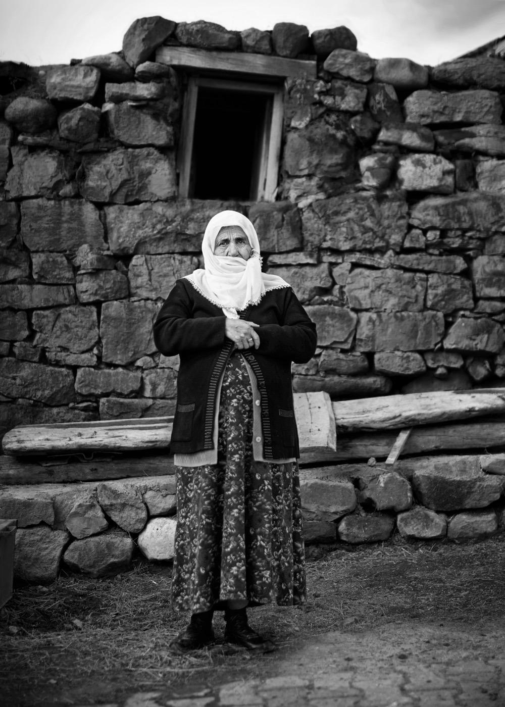 An elderly woman stands before her hand-laid stone house, hands clasped, eastern Anatolia — fine art black-and-white portrait by Sefa Yamak, Anatolia series.
