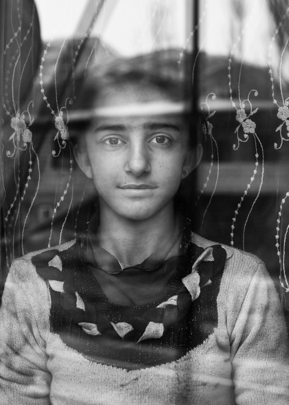 A girl looks out from behind a lace-curtained window, the yard reflected on the glass, eastern Anatolia — fine art black-and-white portrait by Sefa Yamak, Anatolia series.