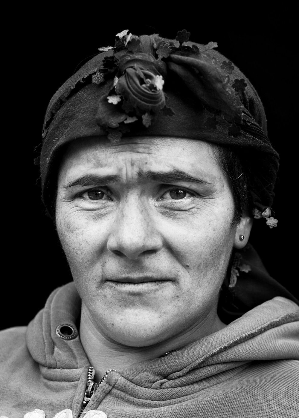A woman in a dark floral scarf and hooded top meets the lens with an unflinching gaze, eastern Anatolia — fine art black-and-white portrait by Sefa Yamak, Anatolia series.