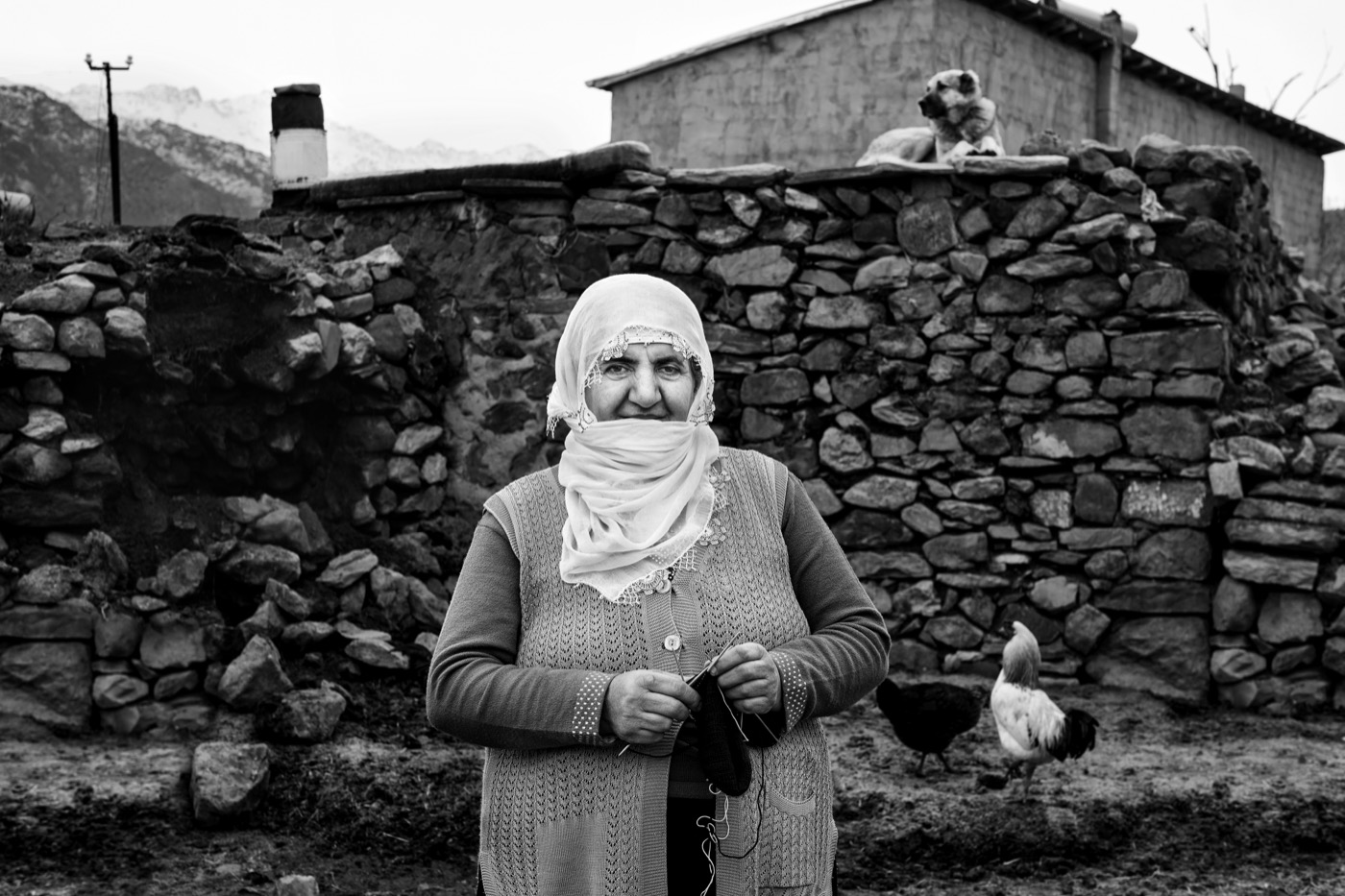 A village woman knits in a muddy yard with chickens and snowy mountains behind, eastern Anatolia — fine art black-and-white portrait by Sefa Yamak, Anatolia series.