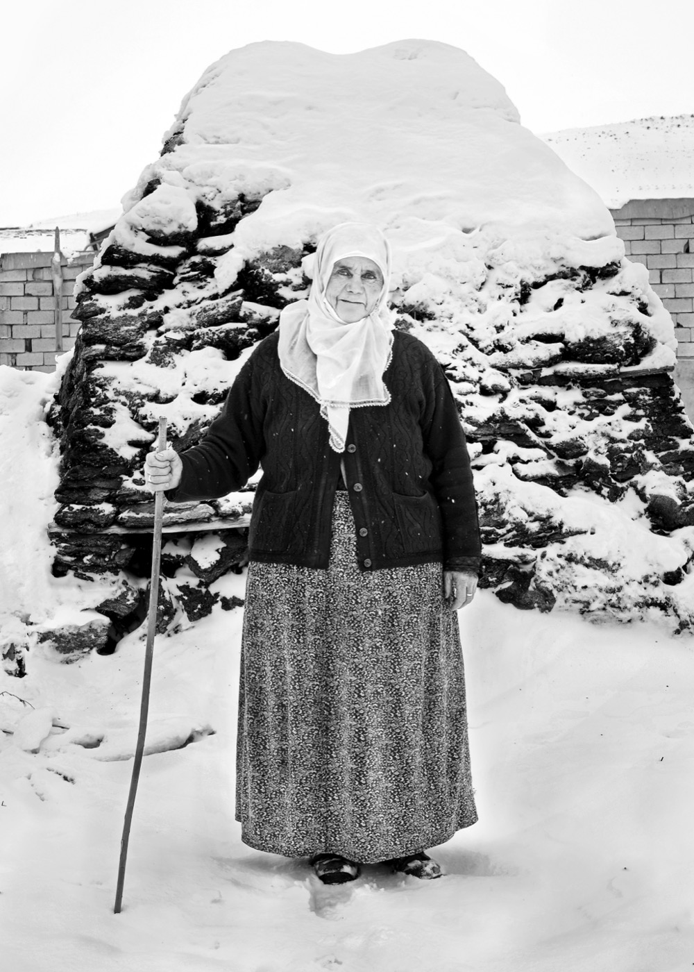 An old woman stands with a cane in deep snow before a snow-capped woodpile, eastern Anatolia — fine art black-and-white portrait by Sefa Yamak, Anatolia series.