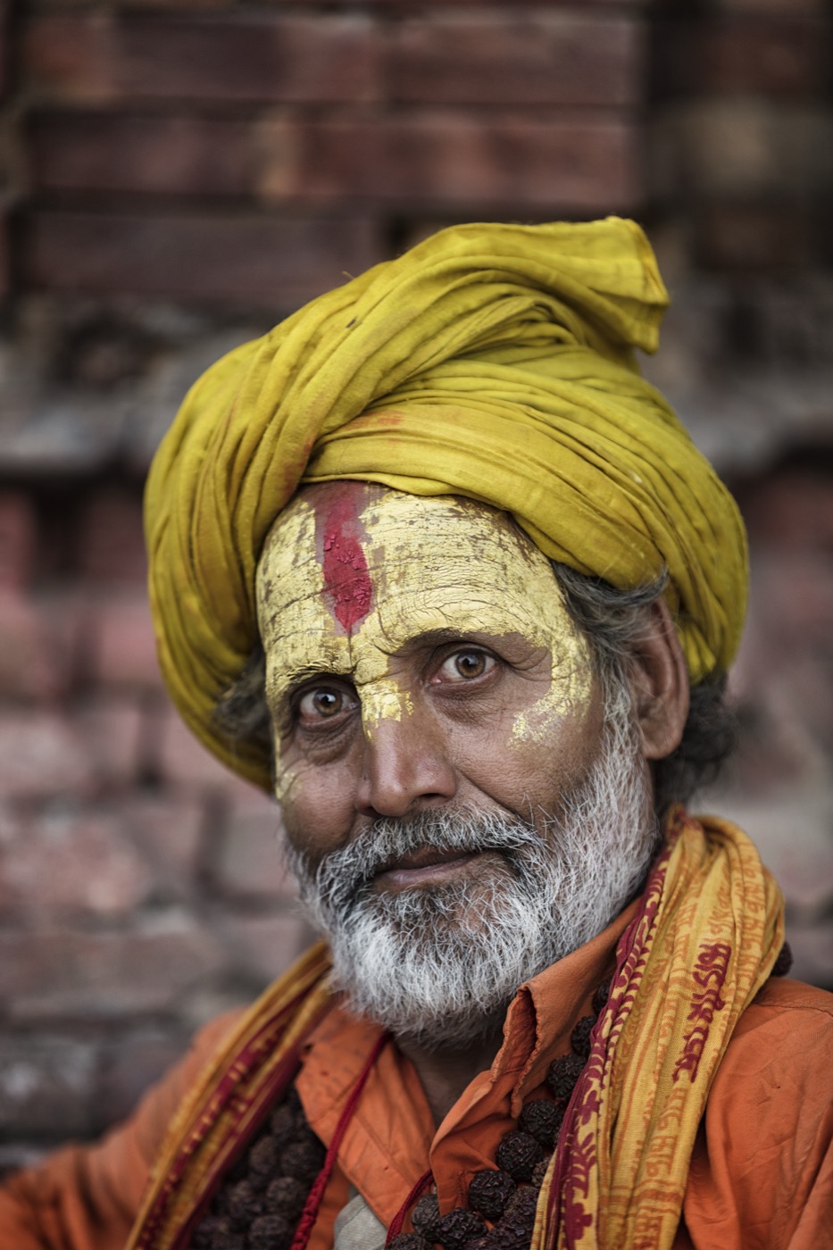 A sadhu in a yellow turban with gold face paint and a red tilak against an old brick wall in Varanasi — fine art portrait by Sefa Yamak, Sadhus series.