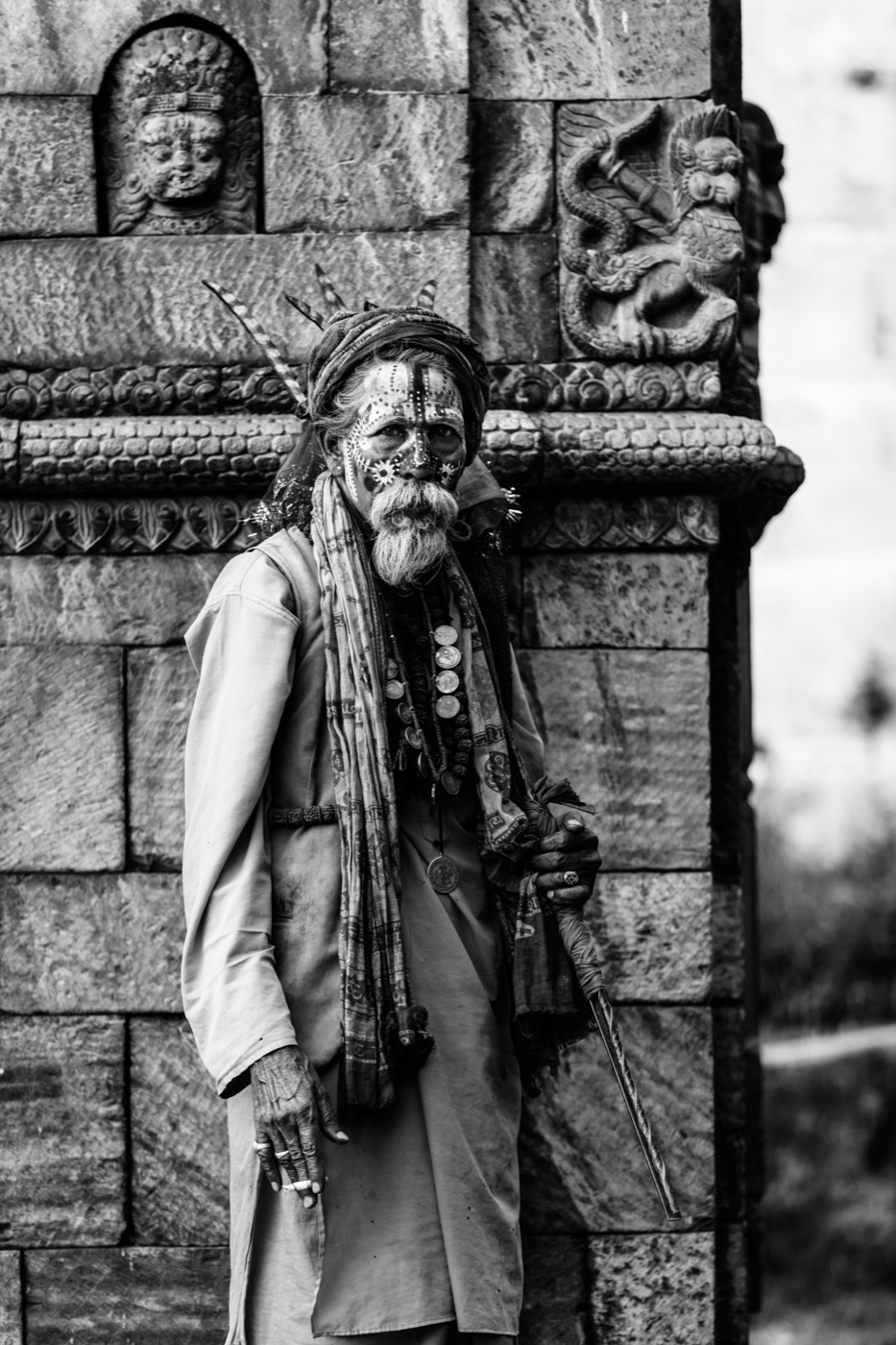 A wandering sadhu with dotted face paint and a staff stands against a carved temple wall — fine art black-and-white portrait by Sefa Yamak, Sadhus series.
