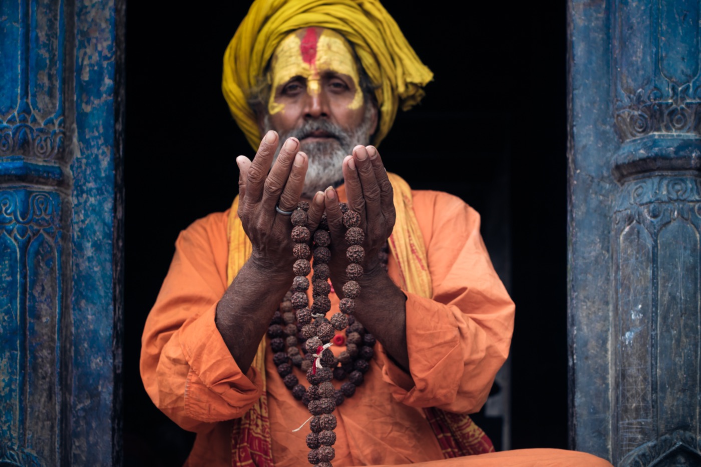 A yellow-turbaned sadhu lifts a rudraksha mala in a weathered blue temple doorway, Varanasi — fine art colour portrait by Sefa Yamak, Sadhus series.